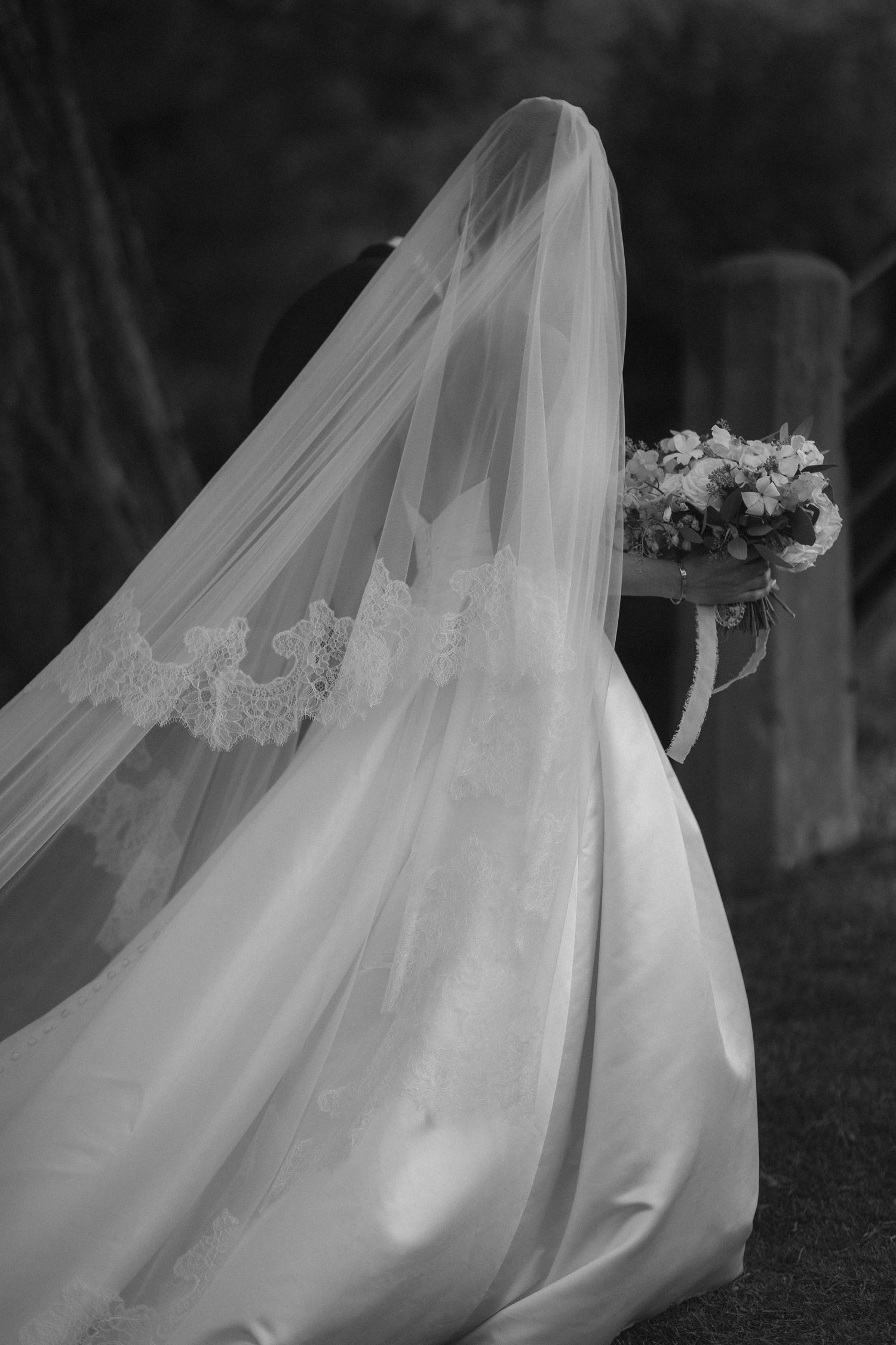 Black and white photo of a bride in a flowing gown with lace-trimmed veil holding a floral bouquet.