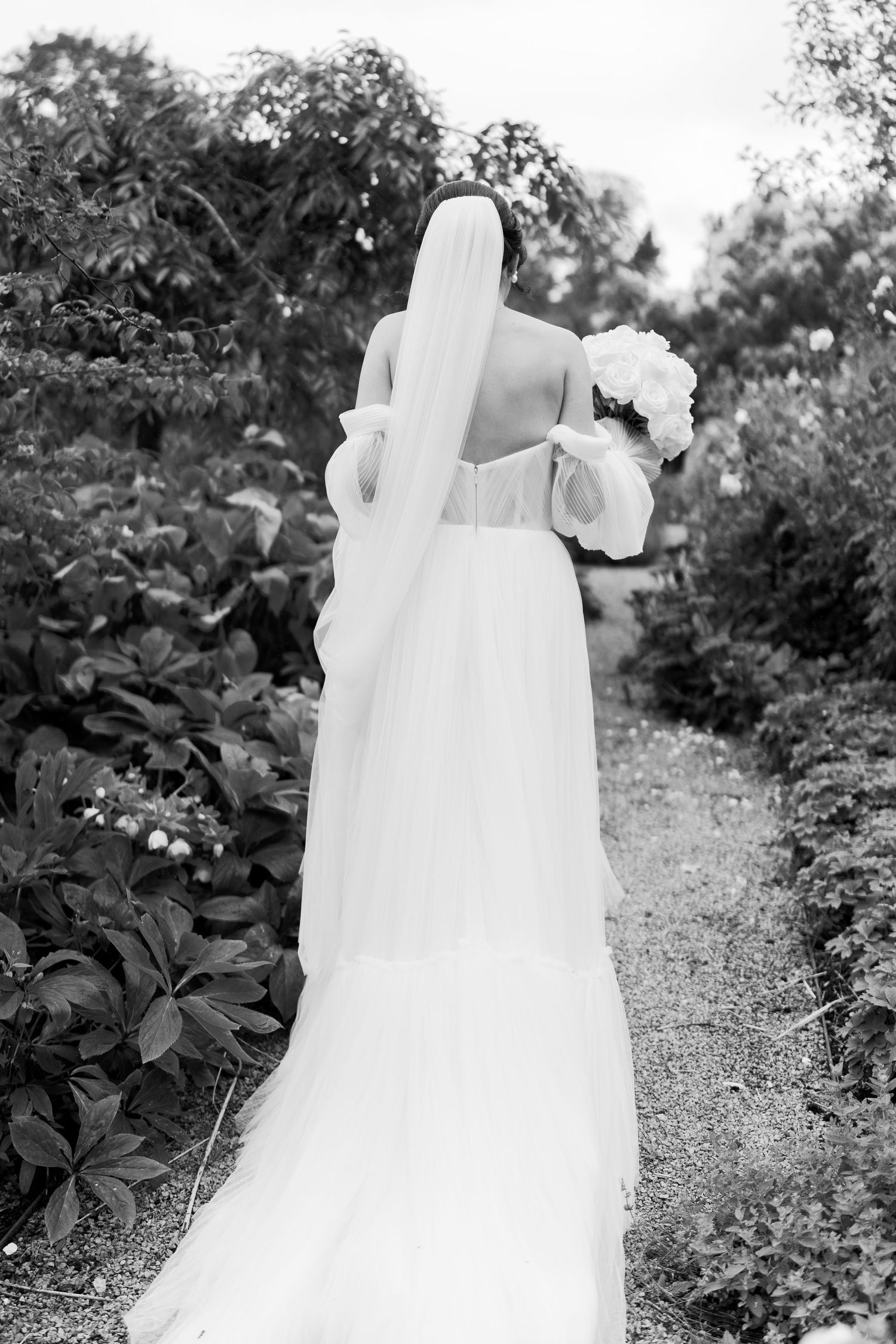 Black and white photo of a bride in a flowing gown and veil, holding a bouquet, walking through a lush garden path.