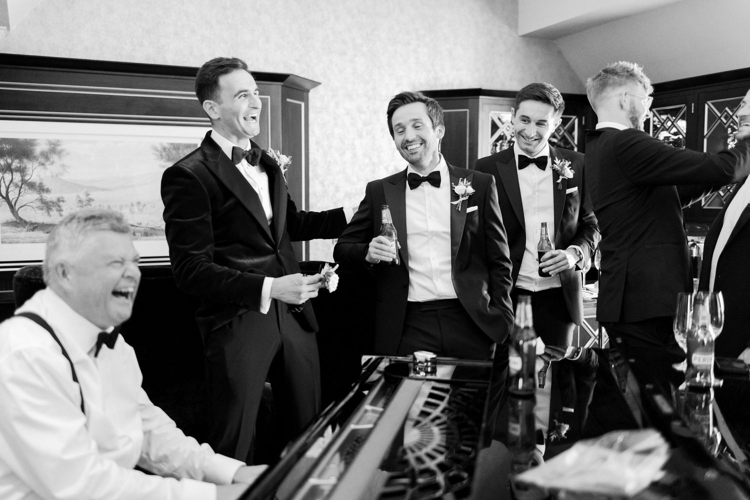 Black and white photo of groomsmen in tuxedos laughing and drinking near a piano at a wedding reception.