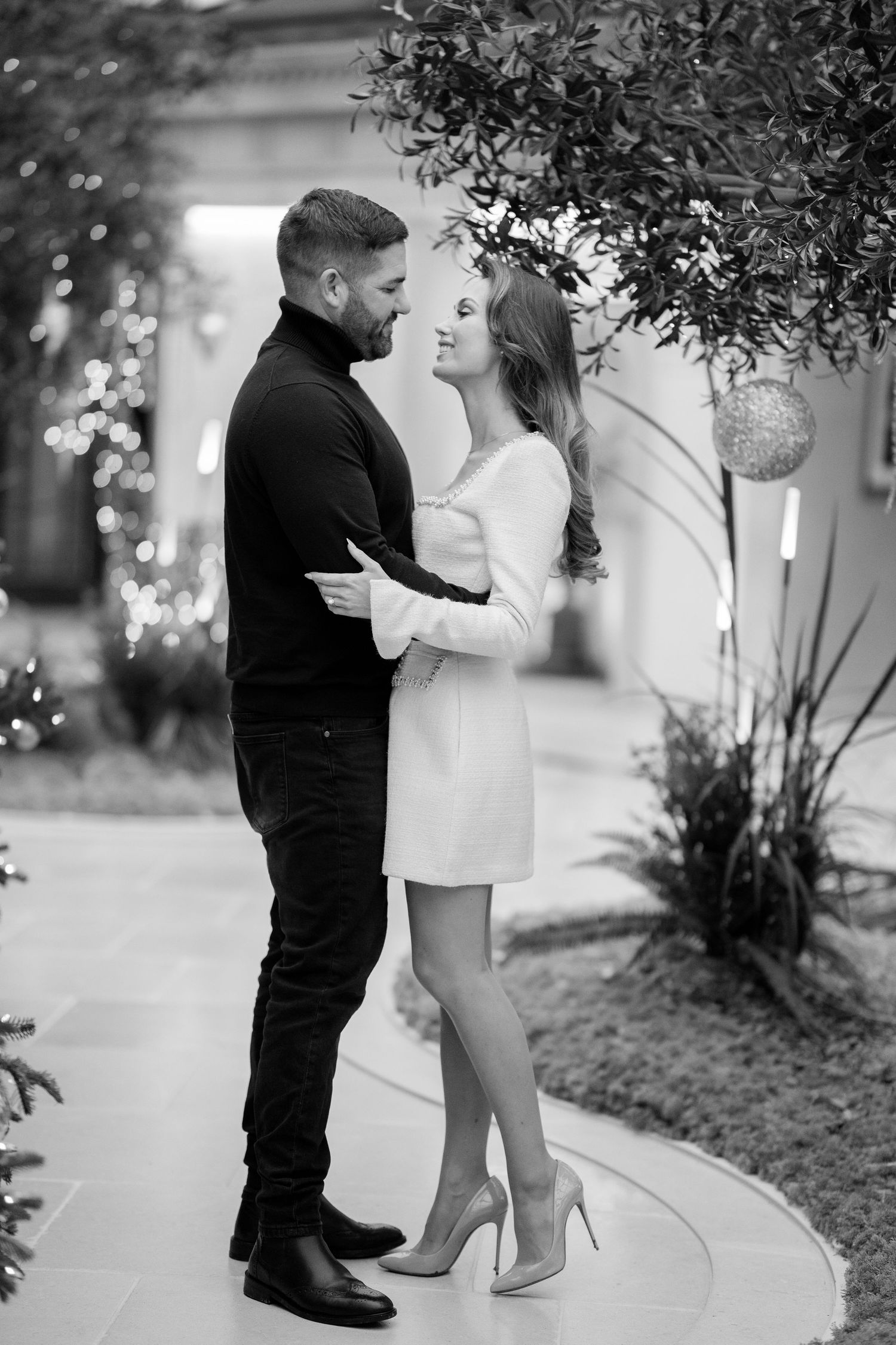 Black and white portrait of stylish couple gazing at each other near olive tree with holiday decorations.