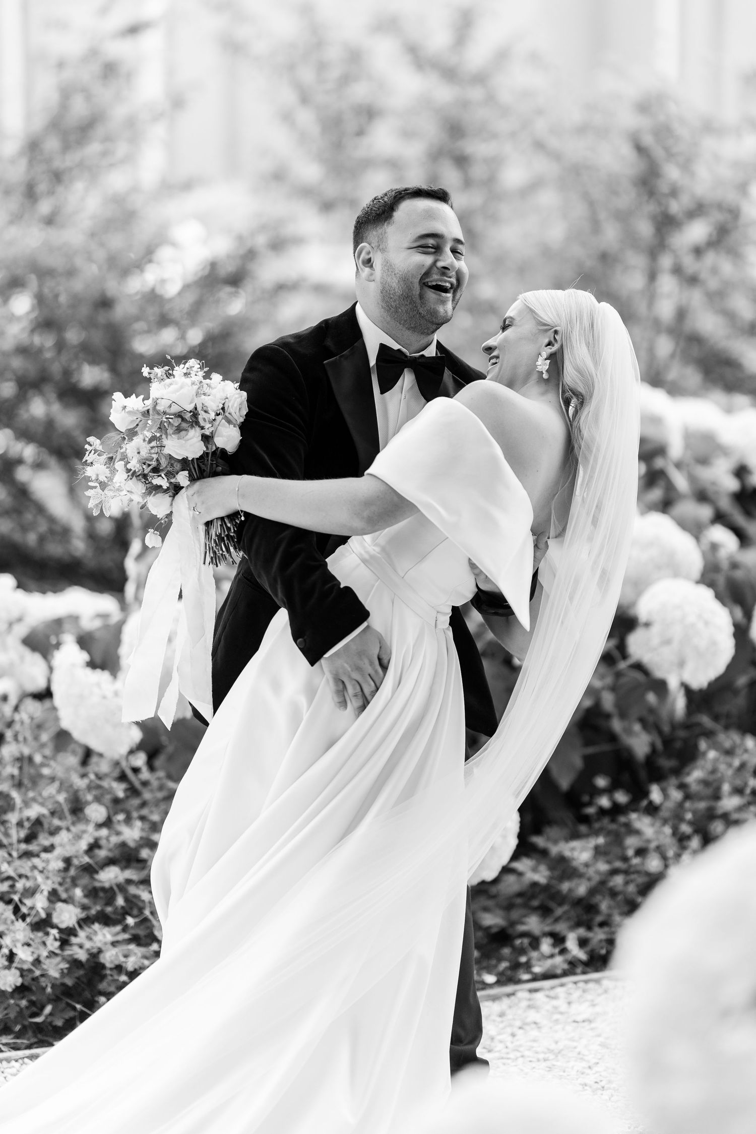 Groom joyfully lifts laughing bride in black-and-white photo among blooming garden flowers.