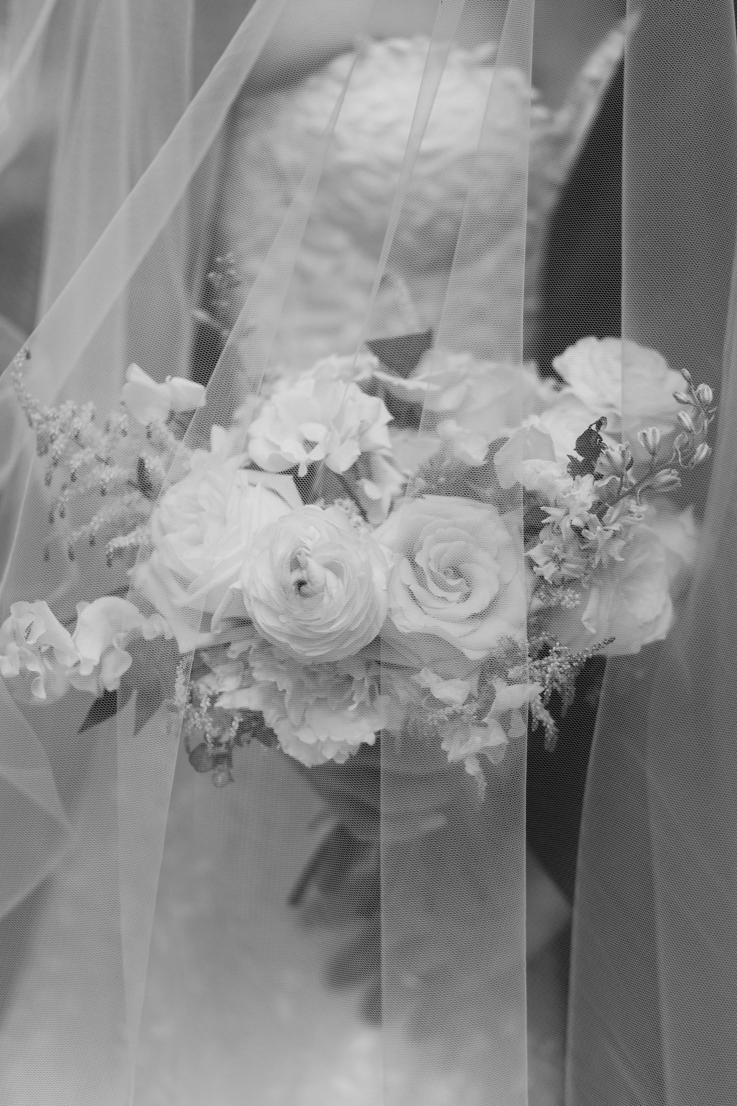 Black and white photo of a bride holding a lush bouquet of roses and flowers, with a flowing veil draped around her.