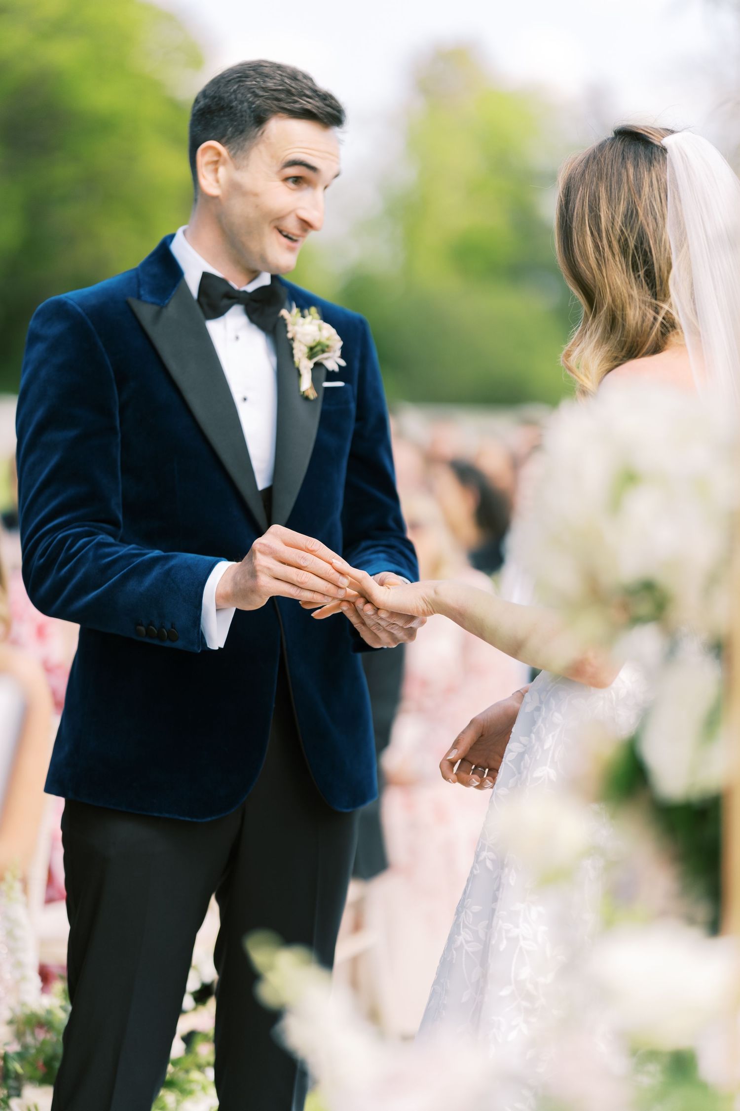 Groom in navy velvet tuxedo places wedding ring on brides finger during outdoor ceremony.