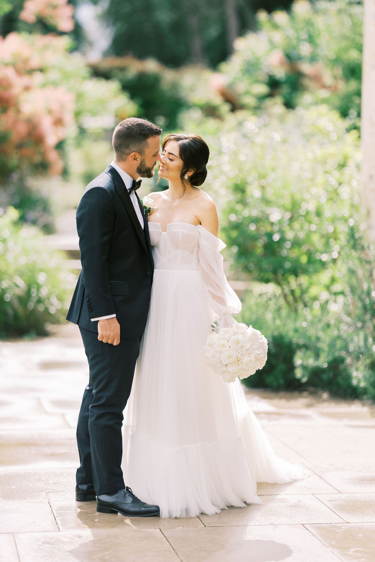 Bride and groom share a romantic moment in a lush garden, bride holding white bouquet in elegant off-shoulder gown.