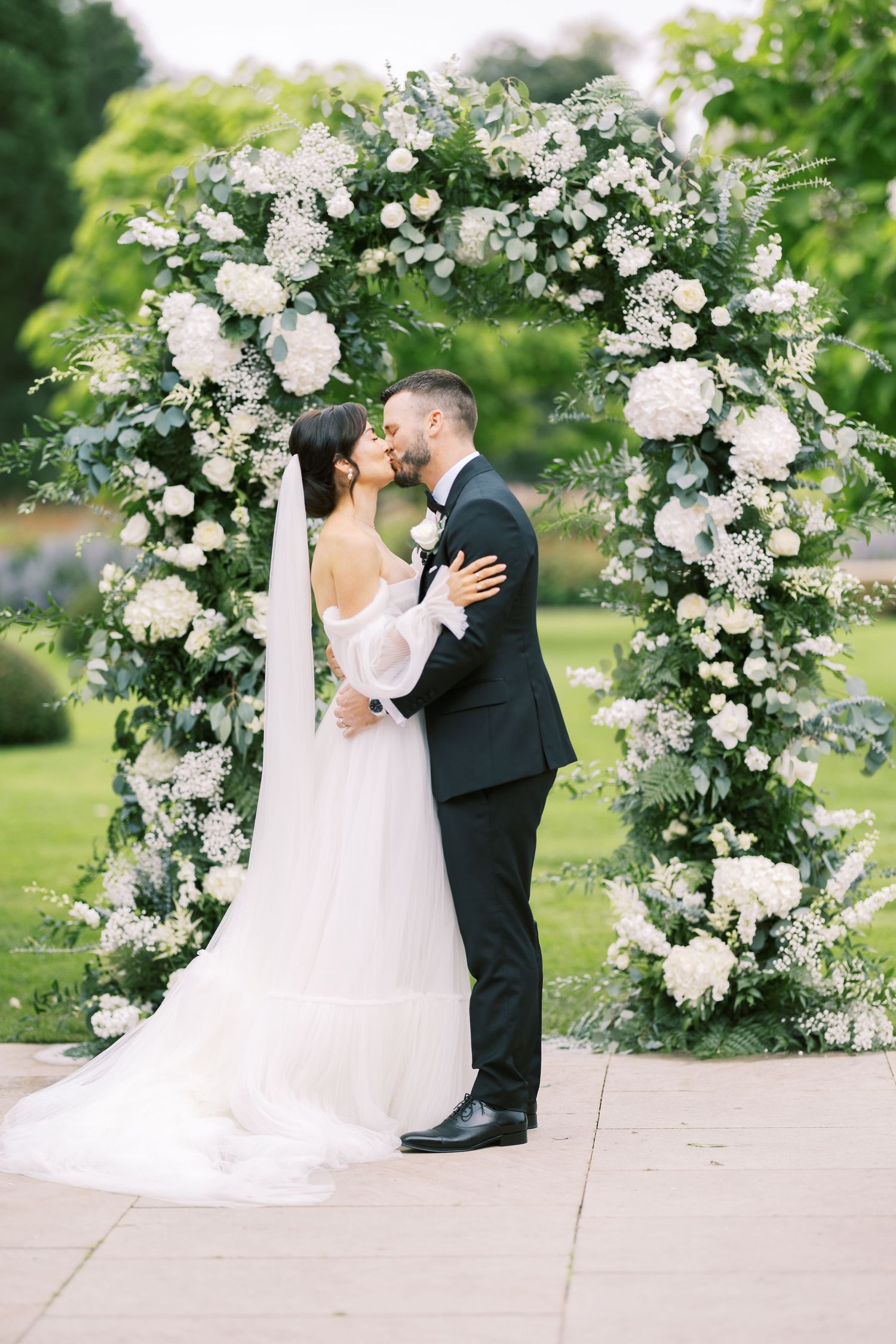 Newlyweds share first kiss under stunning circular white floral arch in lush garden wedding ceremony.