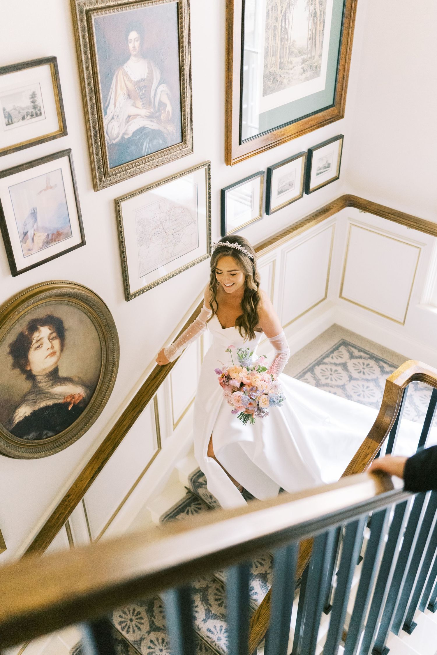 Smiling bride in white gown carrying pastel bouquet ascending grand staircase lined with classic portrait paintings.