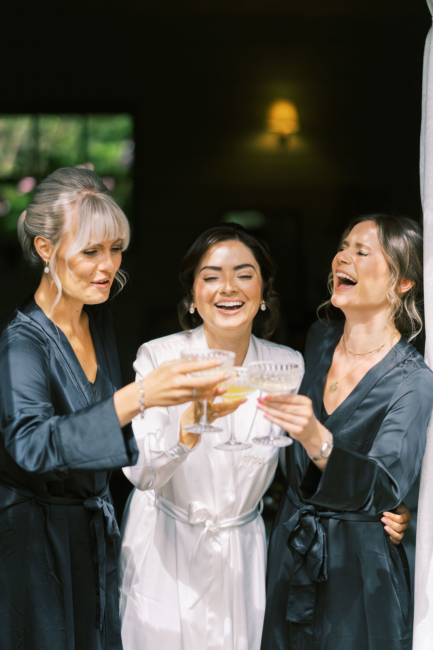 Bride in white robe toasts champagne with two bridesmaids in dark robes, laughing joyfully before the wedding.