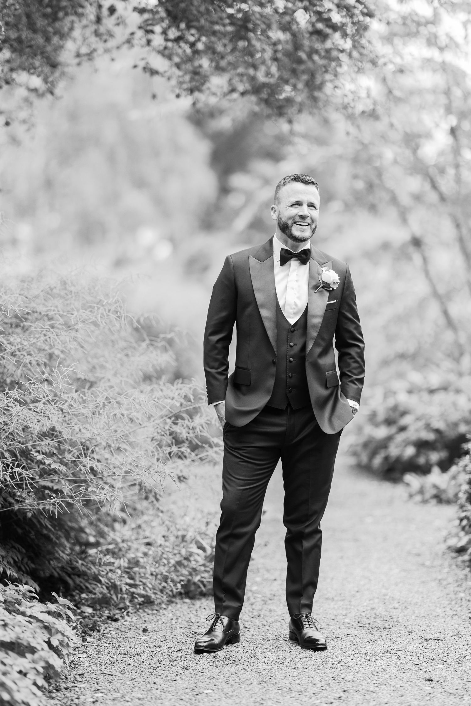 Smiling groom in black tuxedo with boutonniere stands on garden path in elegant black and white wedding portrait.