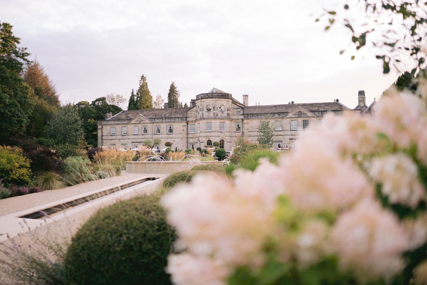 Elegant Georgian manor house surrounded by lush gardens with soft pink blooms in the foreground on a cloudy day.