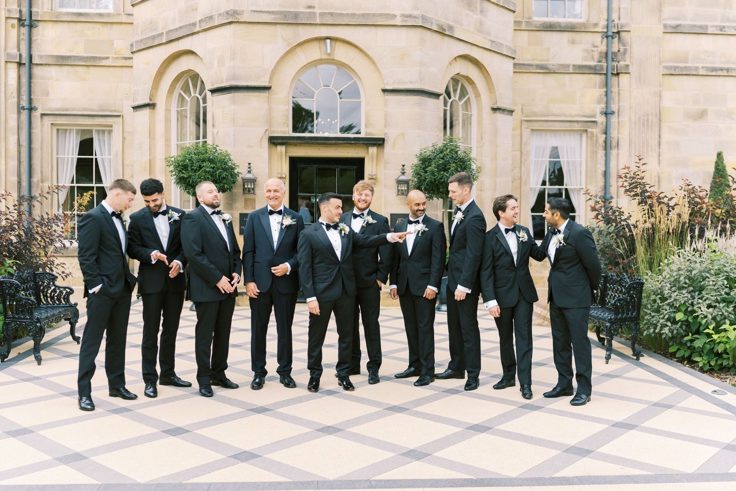 Groomsmen in black tuxedos with bow ties stand together outside an elegant stone building.