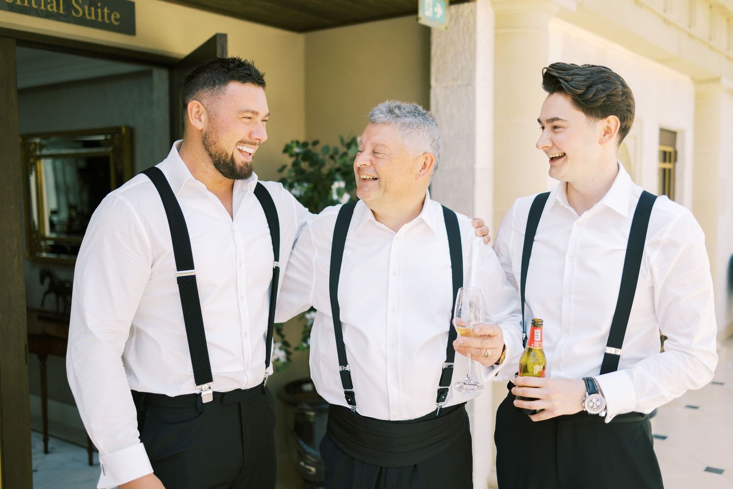 Three men in white shirts and black suspenders smile and celebrate together outside a bridal suite.