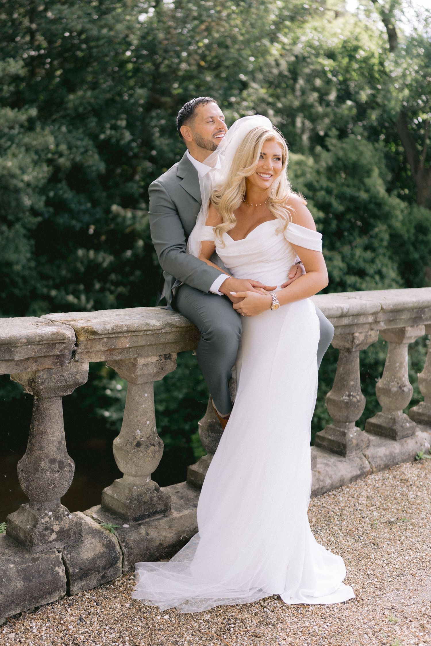 Bride in off-shoulder white gown and groom in gray suit pose on stone balustrade surrounded by lush greenery.