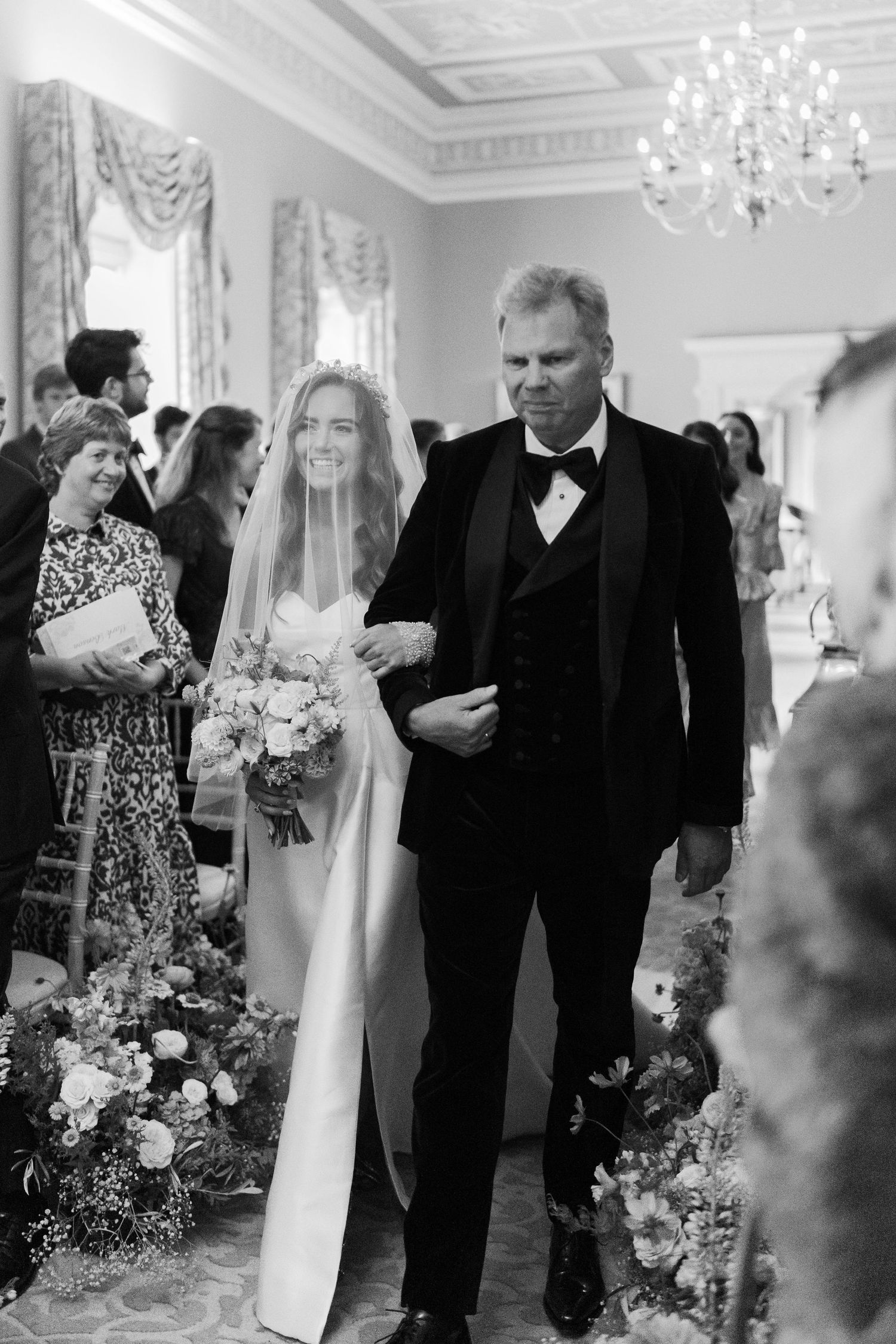 Bride walks down aisle with father in black tuxedo during elegant indoor wedding ceremony.