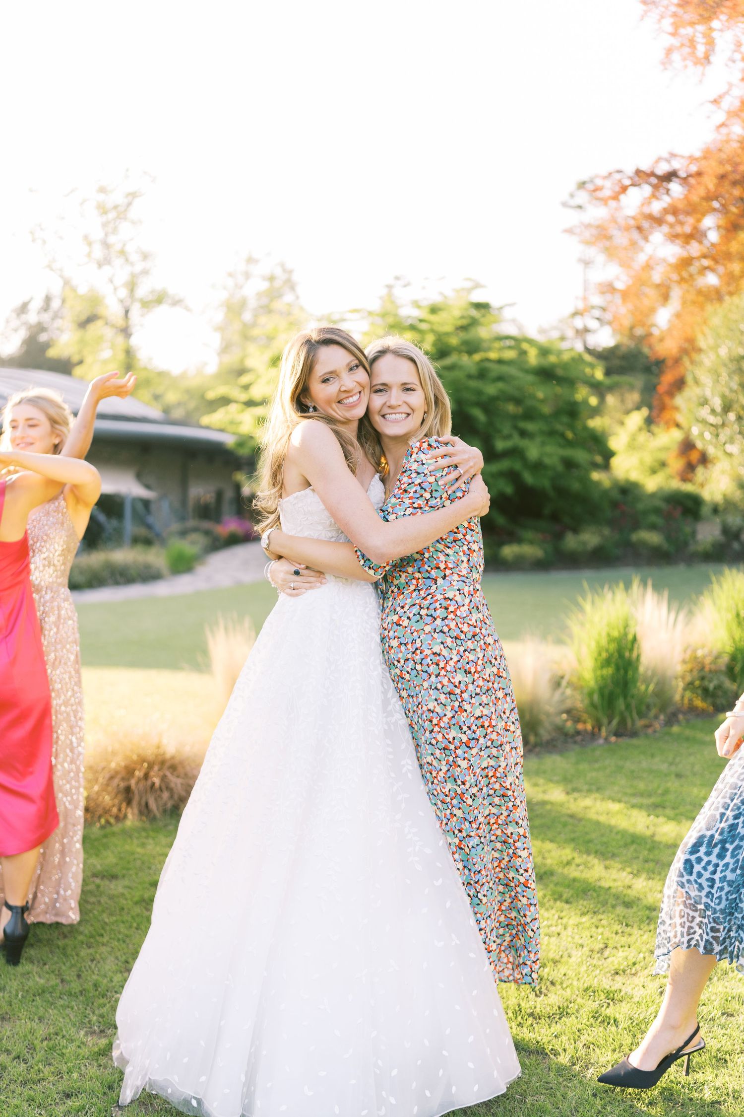 Bride in white ball gown hugging a guest in floral dress at an outdoor wedding reception on a sunny day.