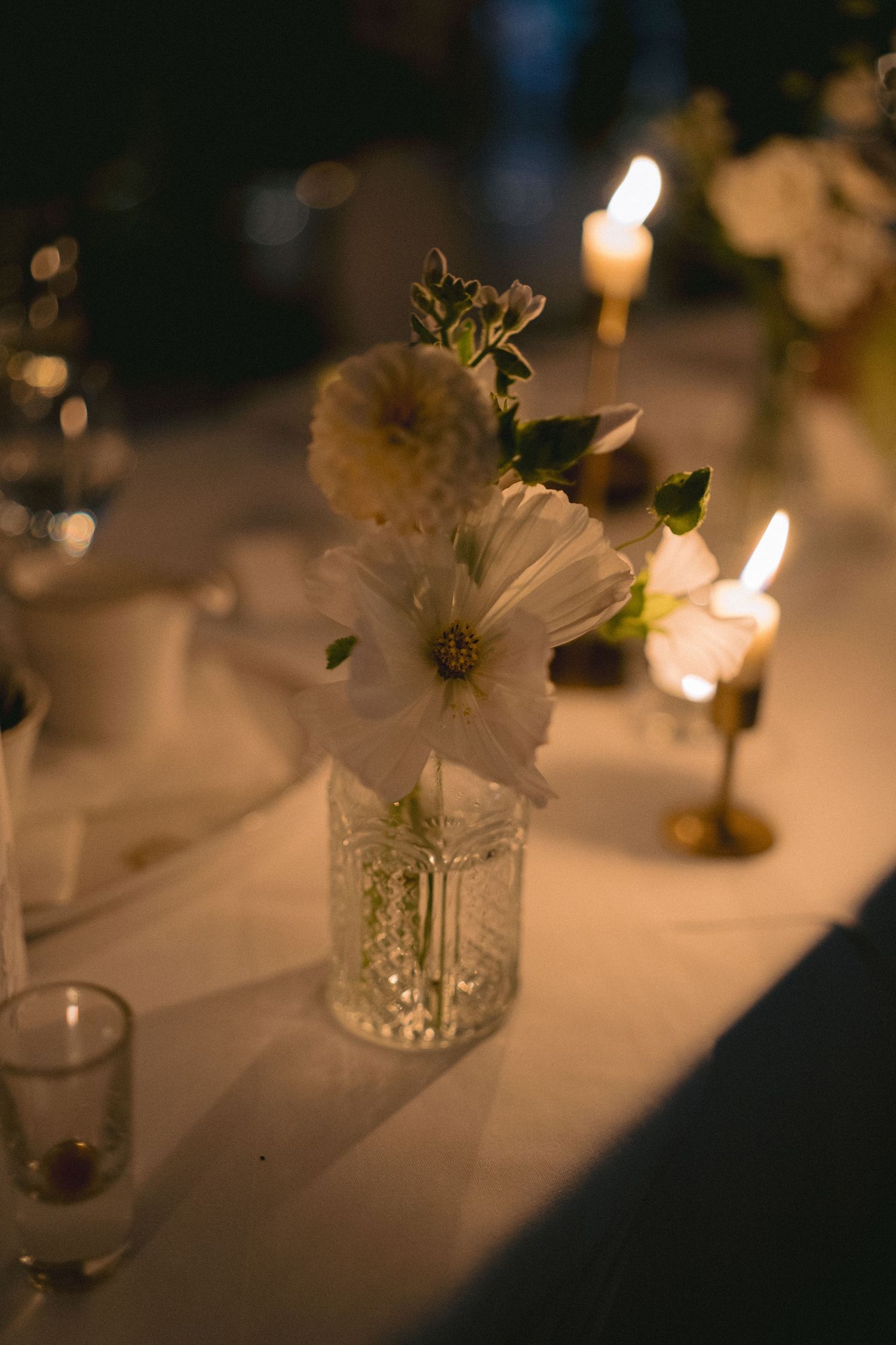White flowers in crystal vase with glowing candles on white tablecloth at elegant dinner setting.