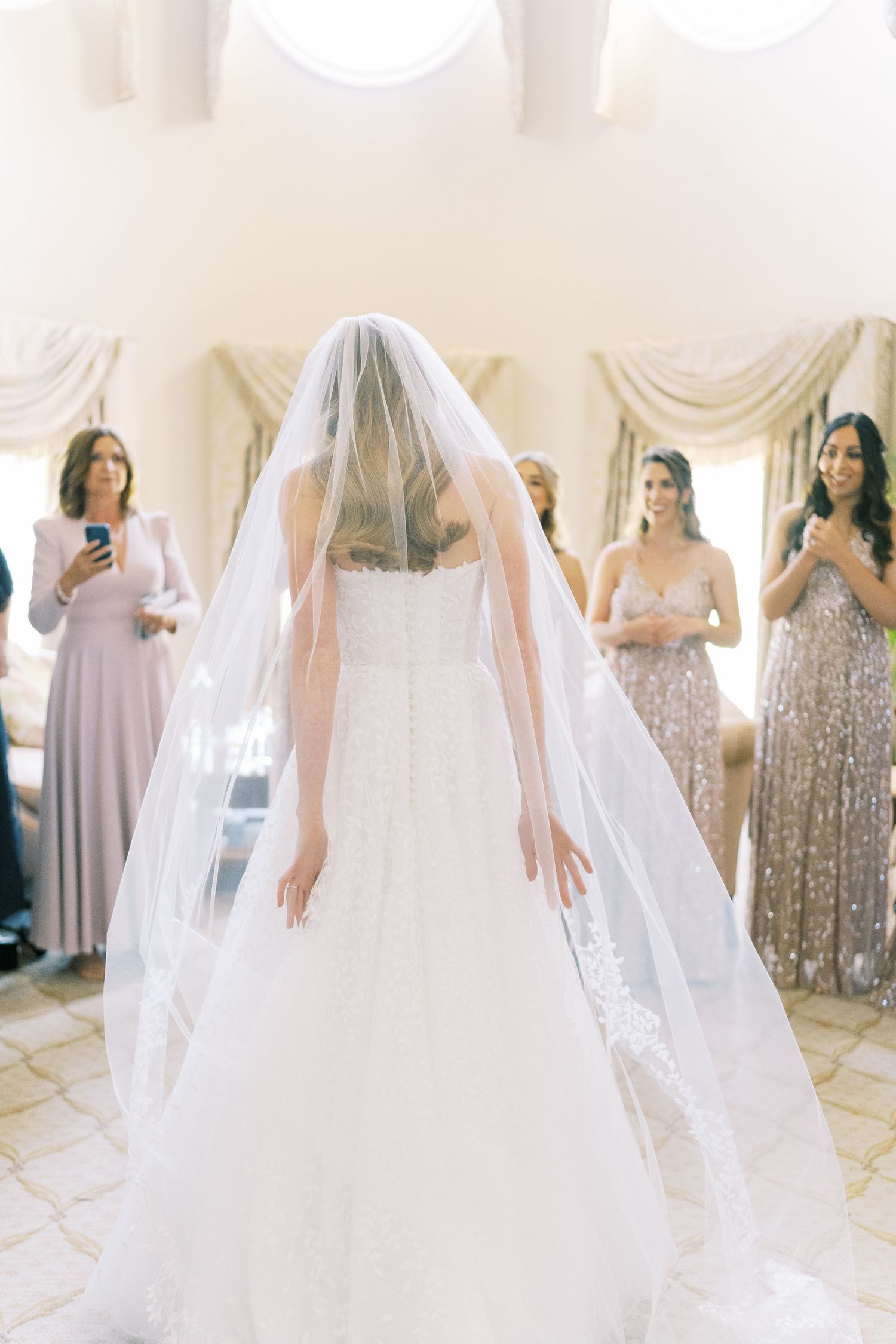 Bride in lace gown and veil revealed to bridesmaids in sparkly dresses during wedding morning prep.