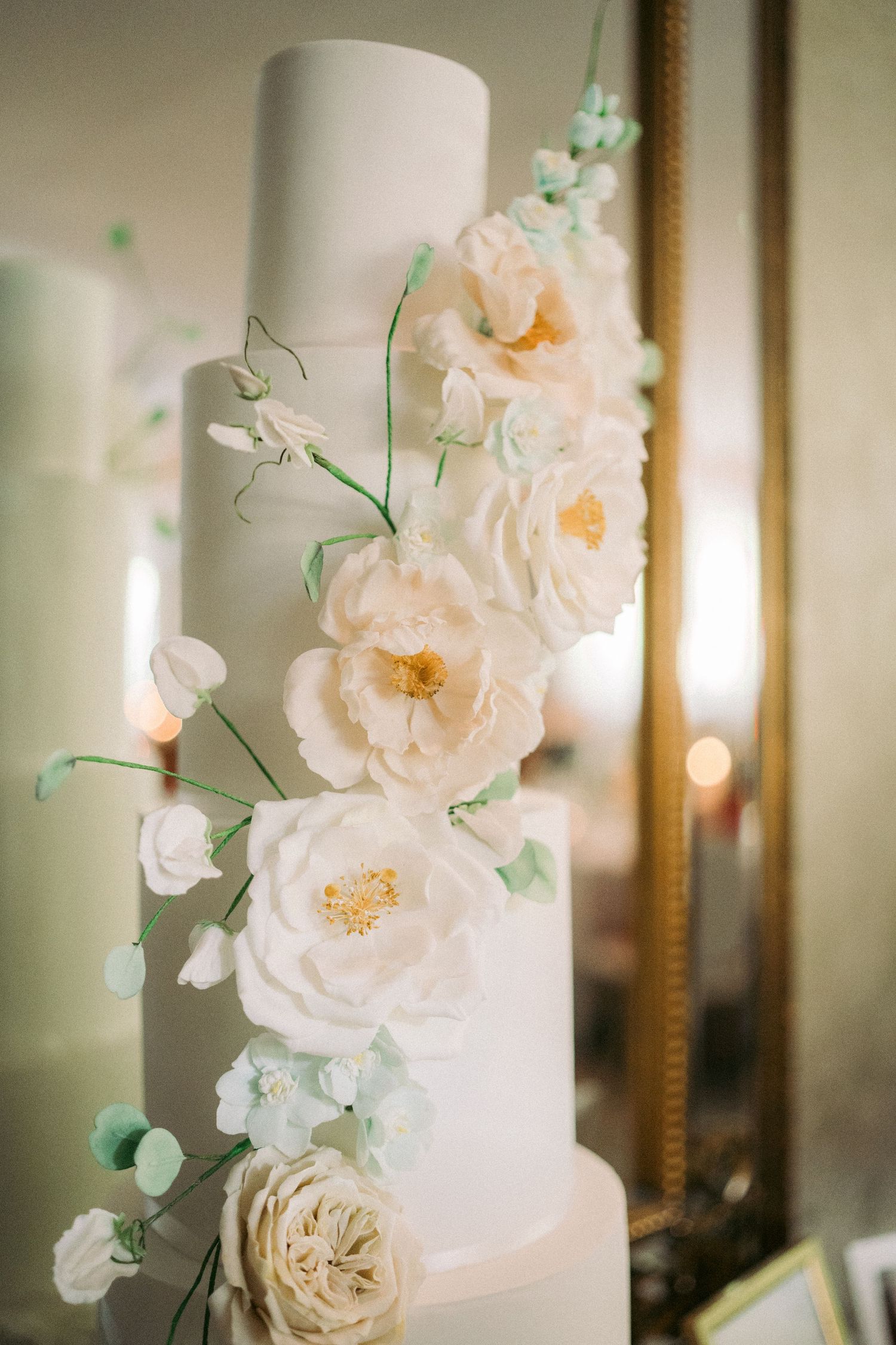 Elegant white wedding cake with delicate sugar flowers in blush and mint tones, displayed against a gold mirror.