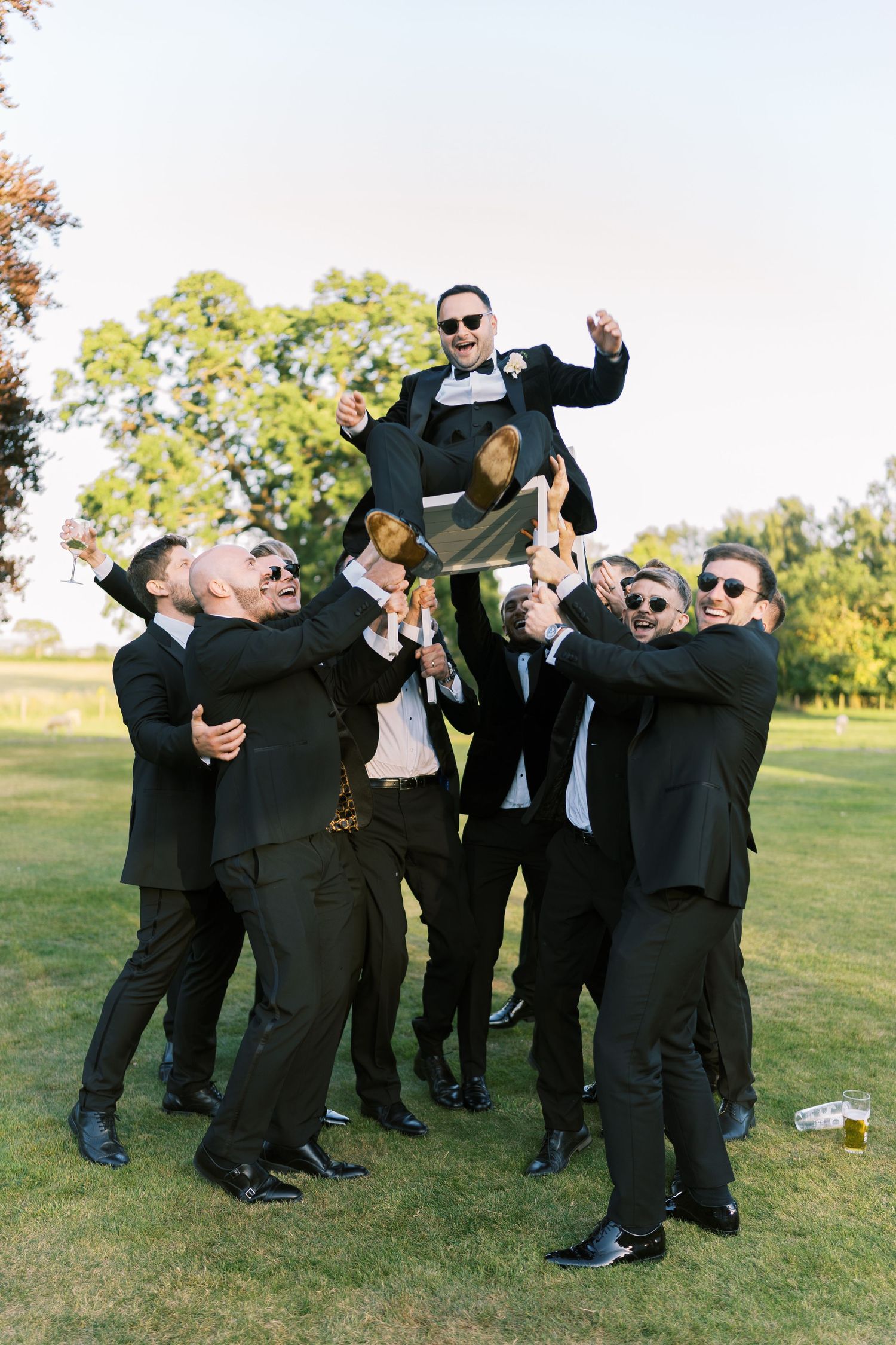 Groomsmen in black suits lifting and celebrating the groom outdoors on a sunny day.