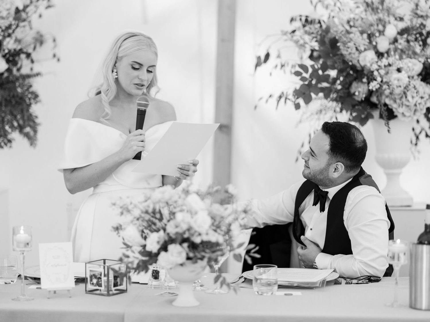 Bride giving heartfelt speech at wedding reception while groom listens adoringly at head table.