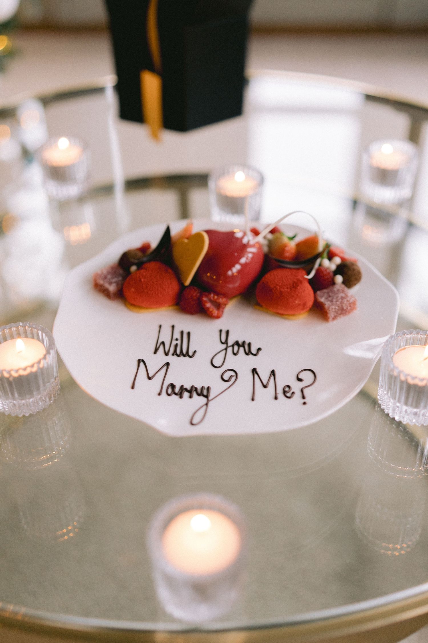 Romantic marriage proposal plate reading Will You Marry Me surrounded by candles and elegant desserts on a glass table.
