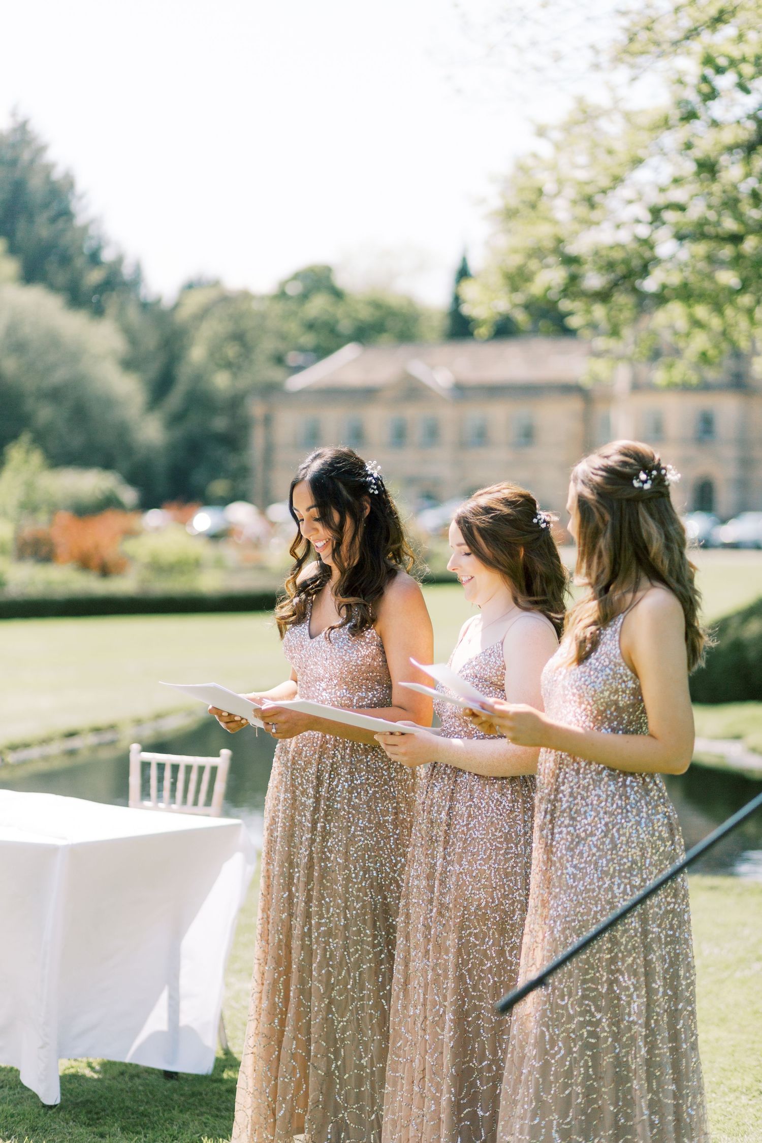 Three bridesmaids in gold sequin gowns reading during outdoor wedding ceremony with manor house in background.