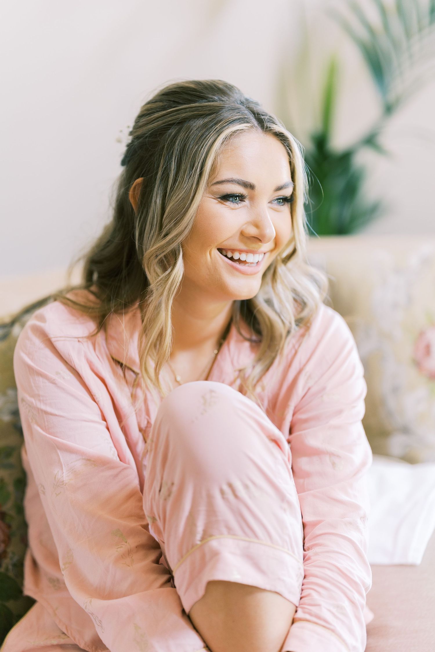 Smiling woman in pink pajamas with wavy blonde hair, sitting relaxed in a bright, cozy room.