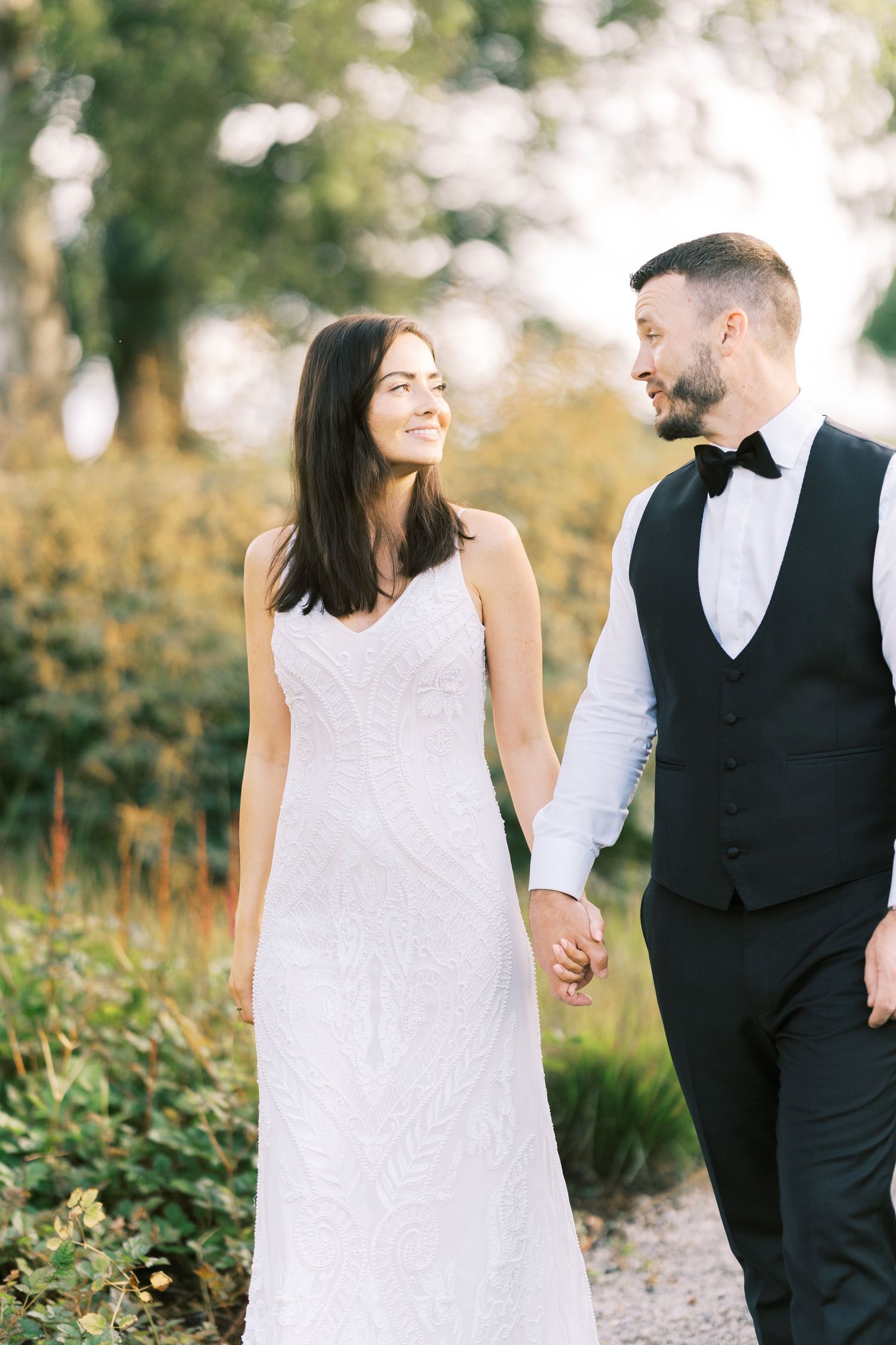 Bride and groom holding hands and smiling while walking outdoors in elegant wedding attire.