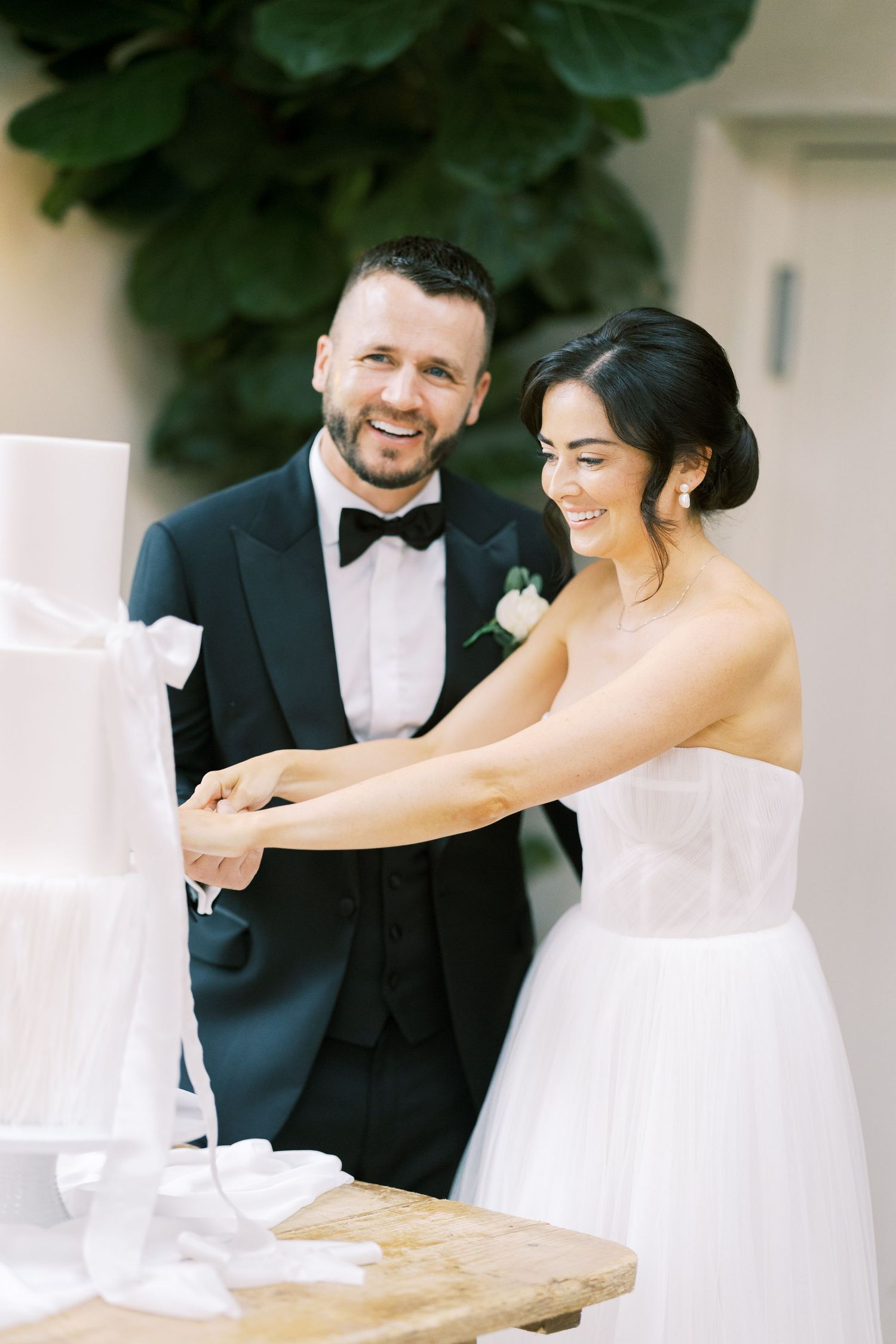 Smiling bride and groom cut elegant white tiered wedding cake adorned with white ribbon bows together.