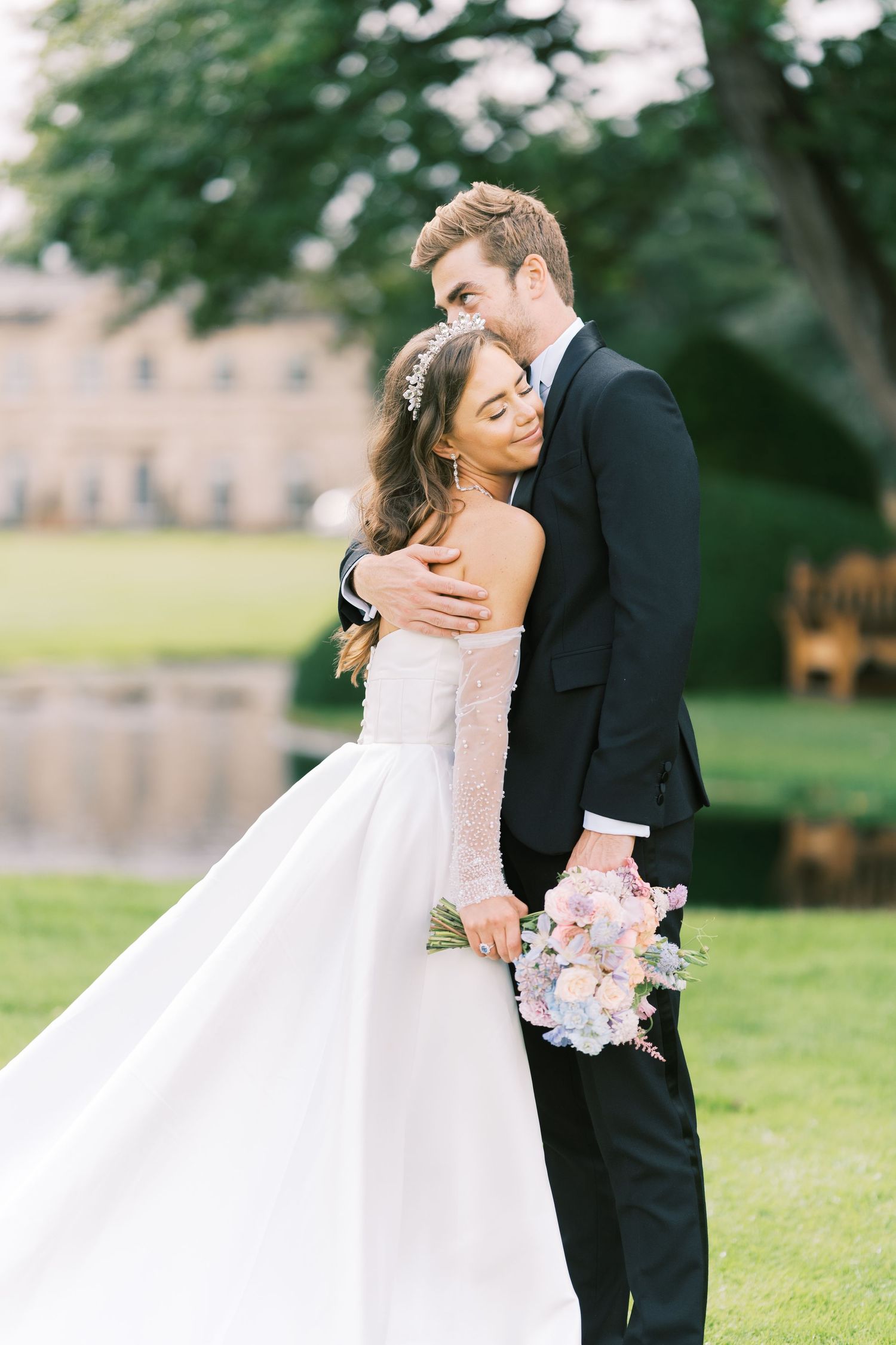 Bride and groom embrace outdoors, she holds pastel bouquet in white gown, he wears black tuxedo, elegant estate backdrop.