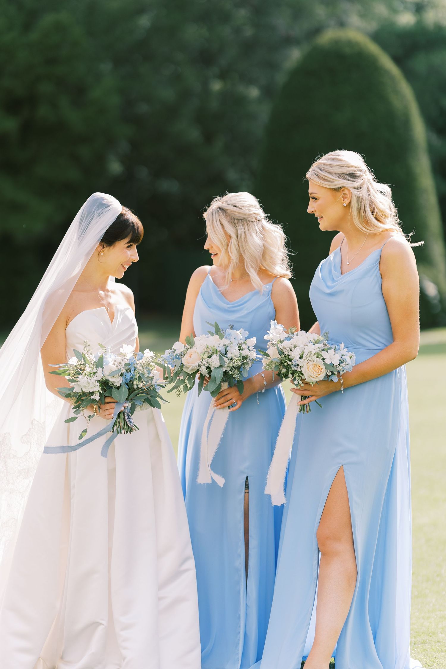 Bride in white gown with veil stands with two bridesmaids in light blue dresses holding floral bouquets outdoors.
