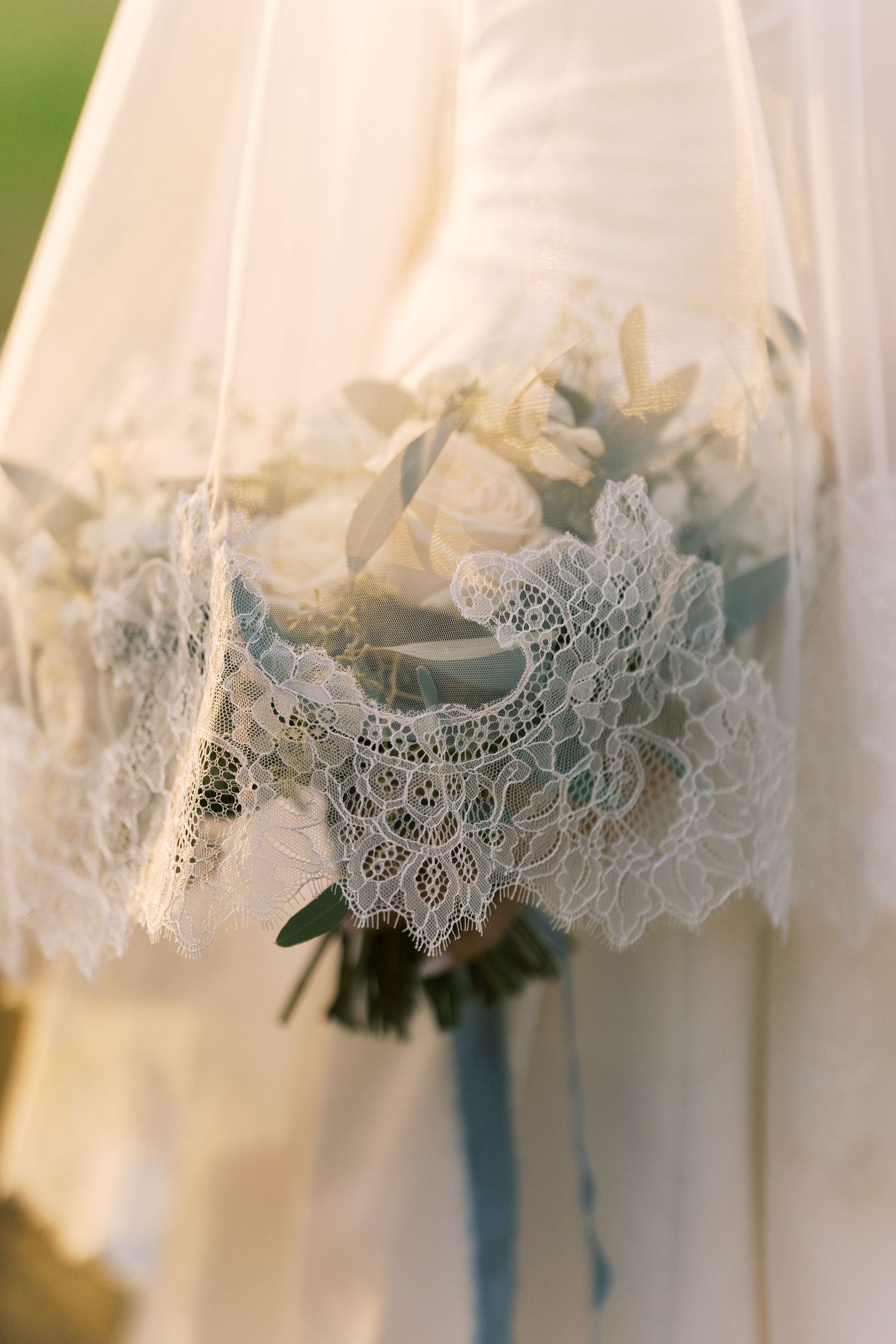 Bride holding a white rose bouquet with lace detail and blue ribbon, wearing an elegant white wedding dress in soft golden light.