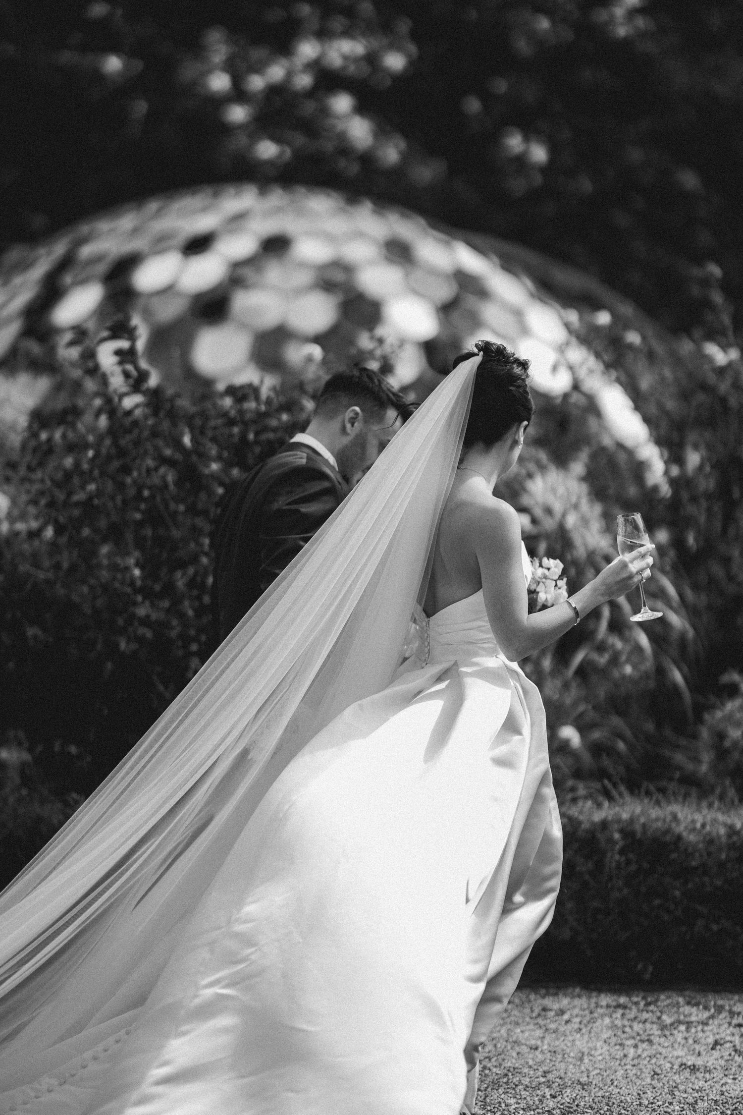 Bride and groom share a moment in garden, her long veil flowing, holding champagne glasses, black and white.