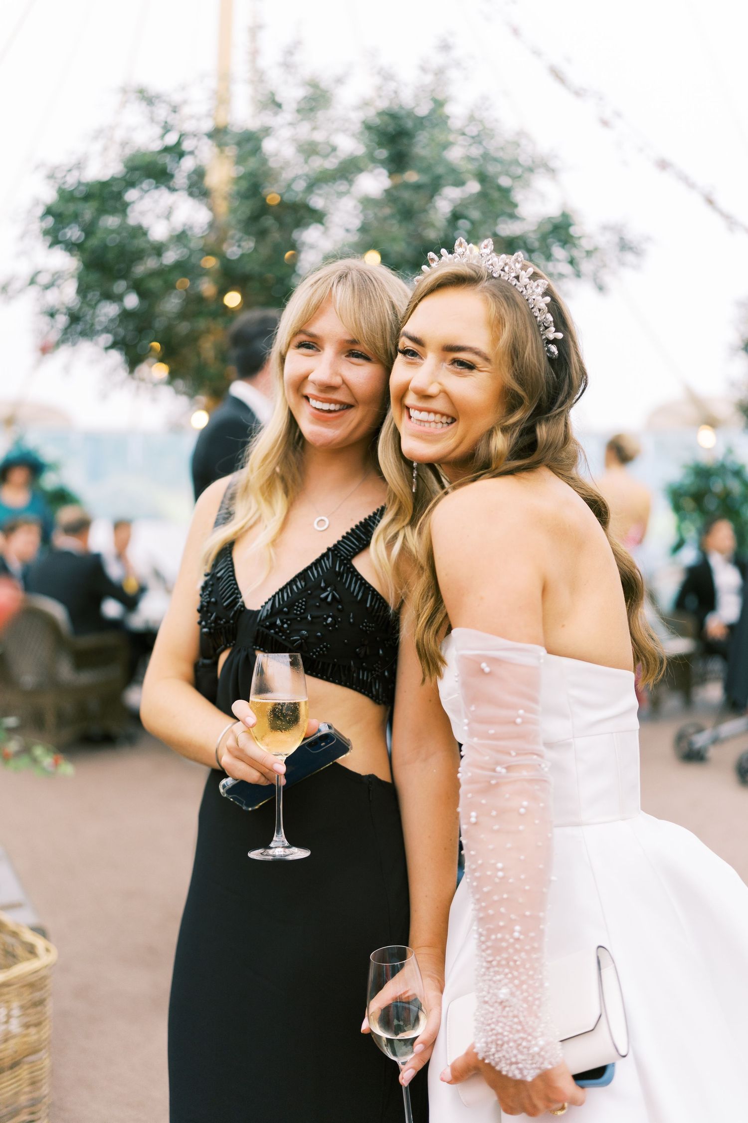 Bride in white strapless gown and tiara smiles with guest in black dress, both holding champagne at wedding reception.