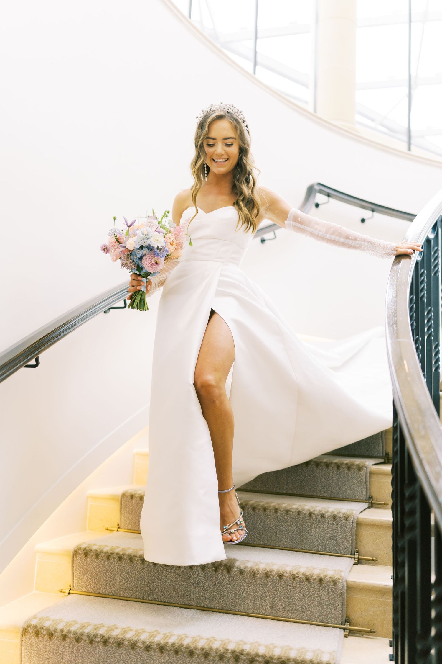 Bride in white gown with slit holds colorful bouquet, descending elegant curved staircase, wearing tiara.