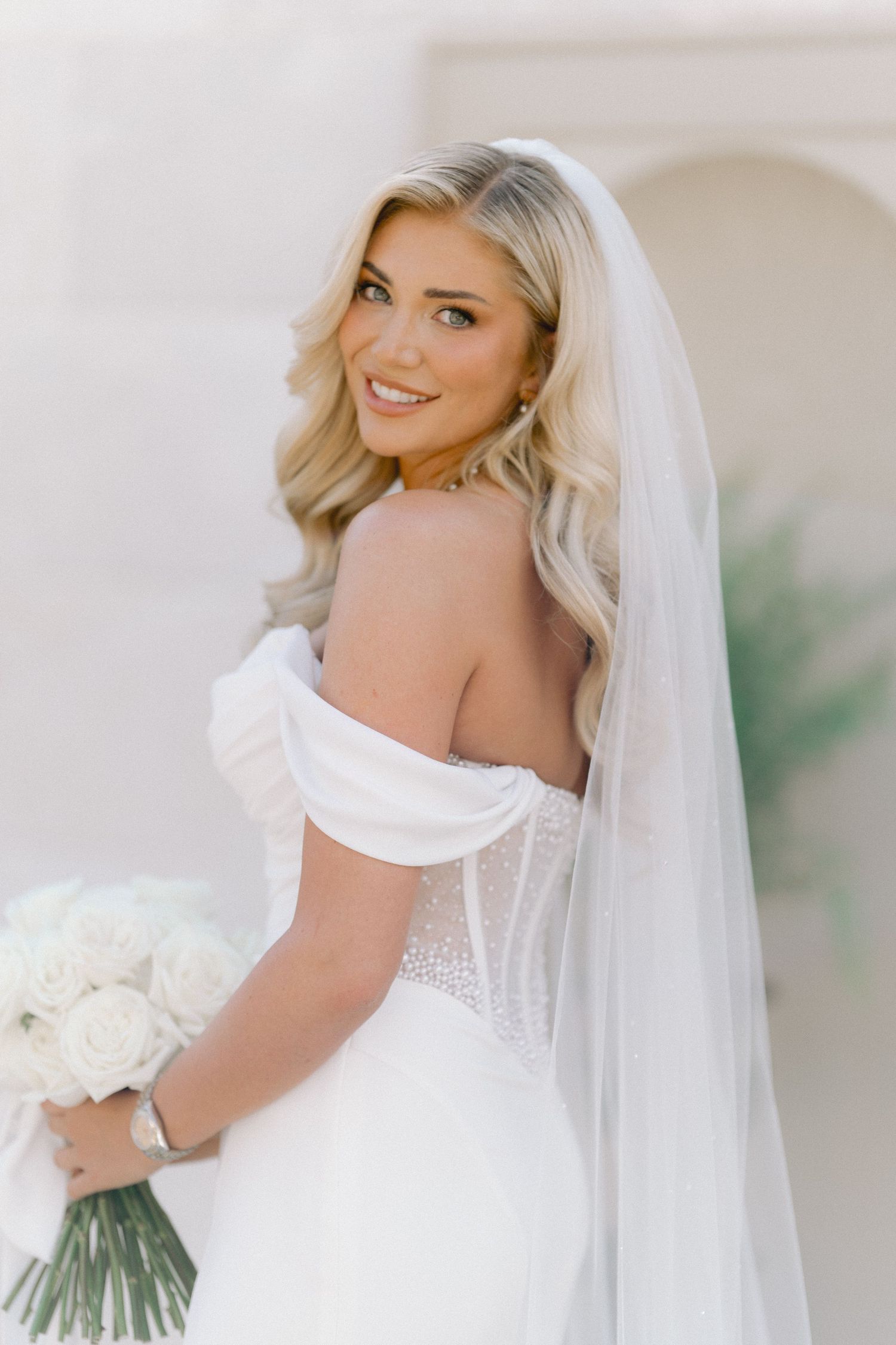 Smiling bride in off-shoulder white gown with veil holds white rose bouquet, posing elegantly for bridal portrait.