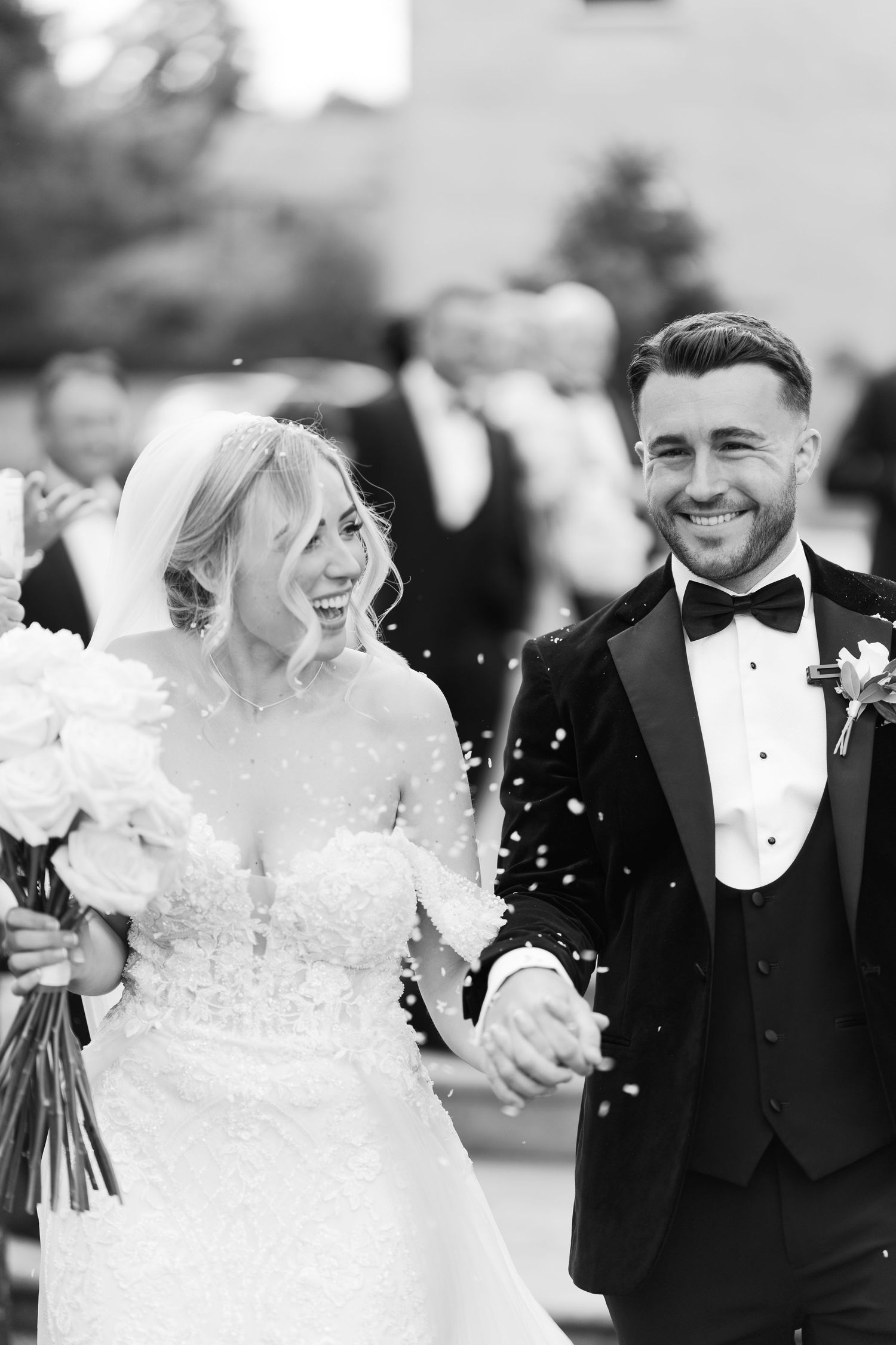Black and white photo of a joyful bride and groom holding hands, laughing as confetti falls during their wedding.