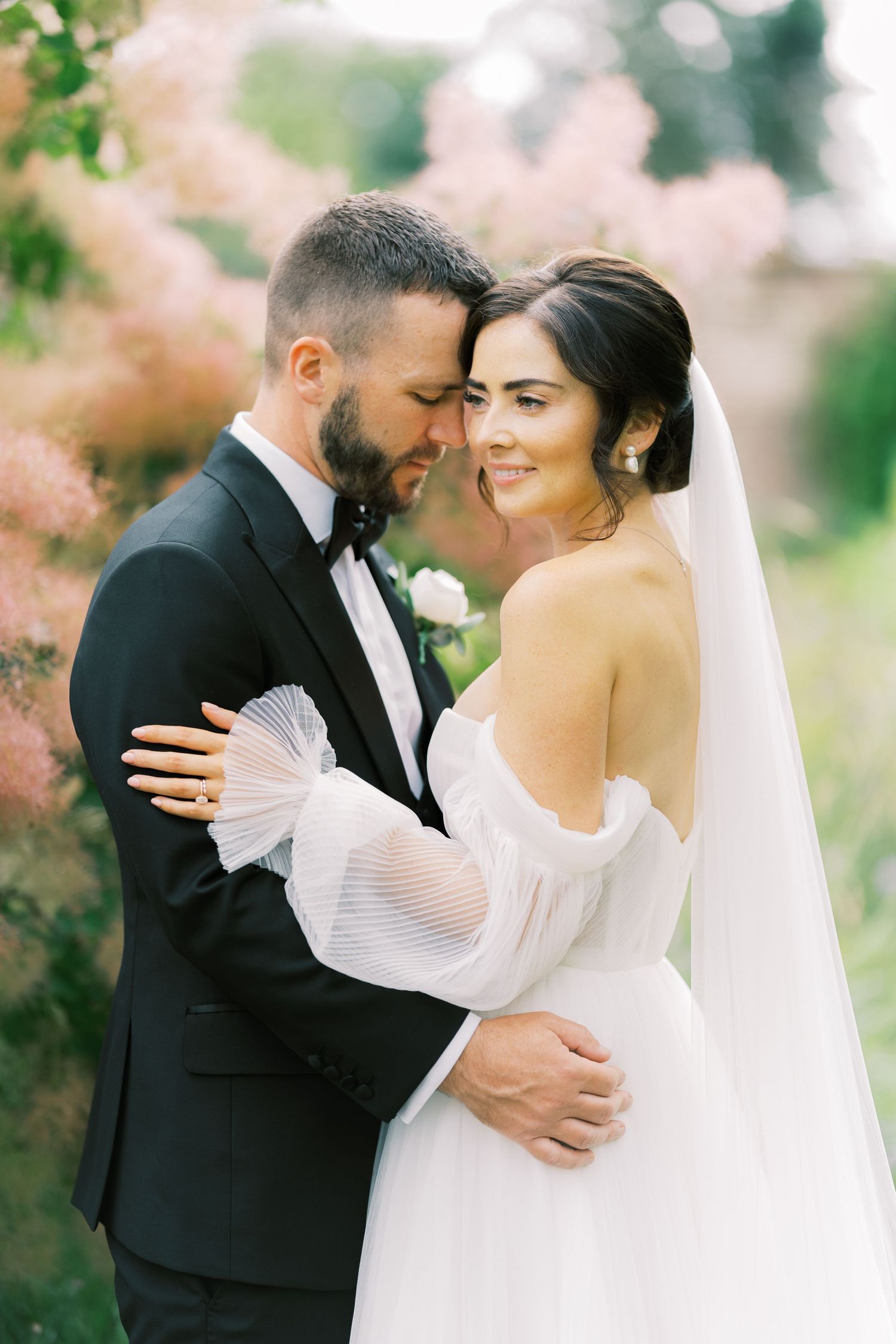 Bride and groom embrace tenderly outdoors with soft pink blooms in the background, bride in off-shoulder gown.