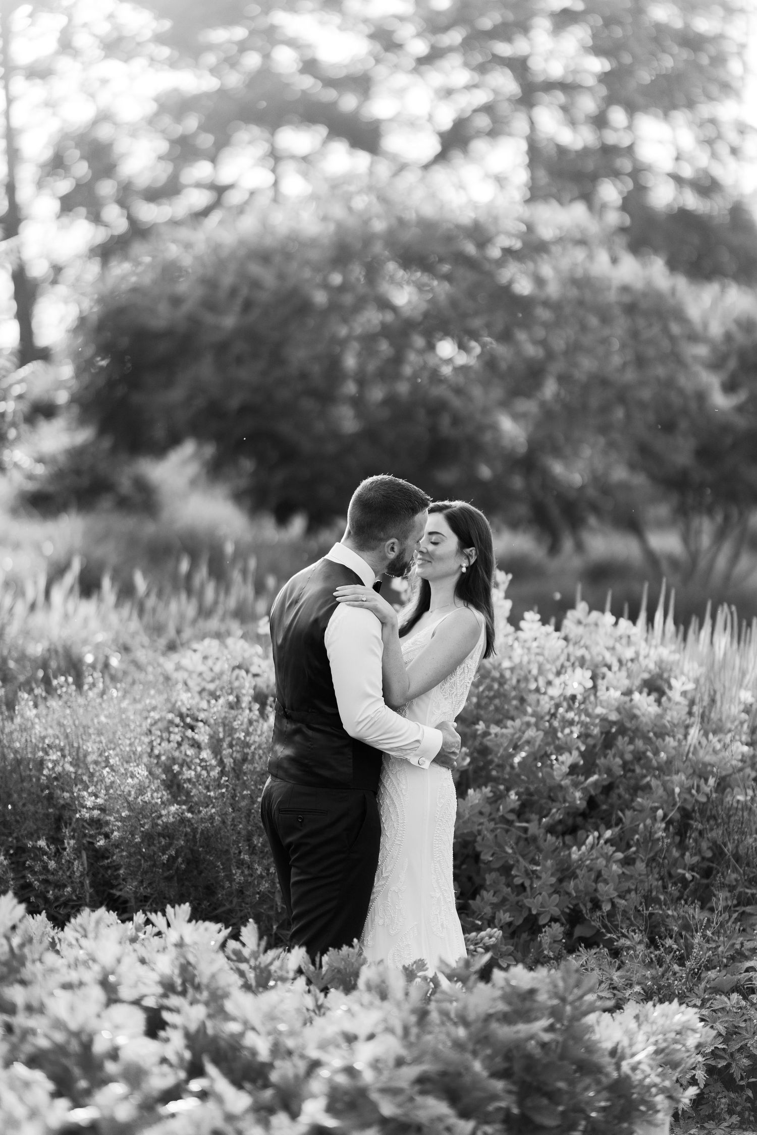 Black and white photo of bride and groom embracing in a lush garden, sharing an intimate moment.