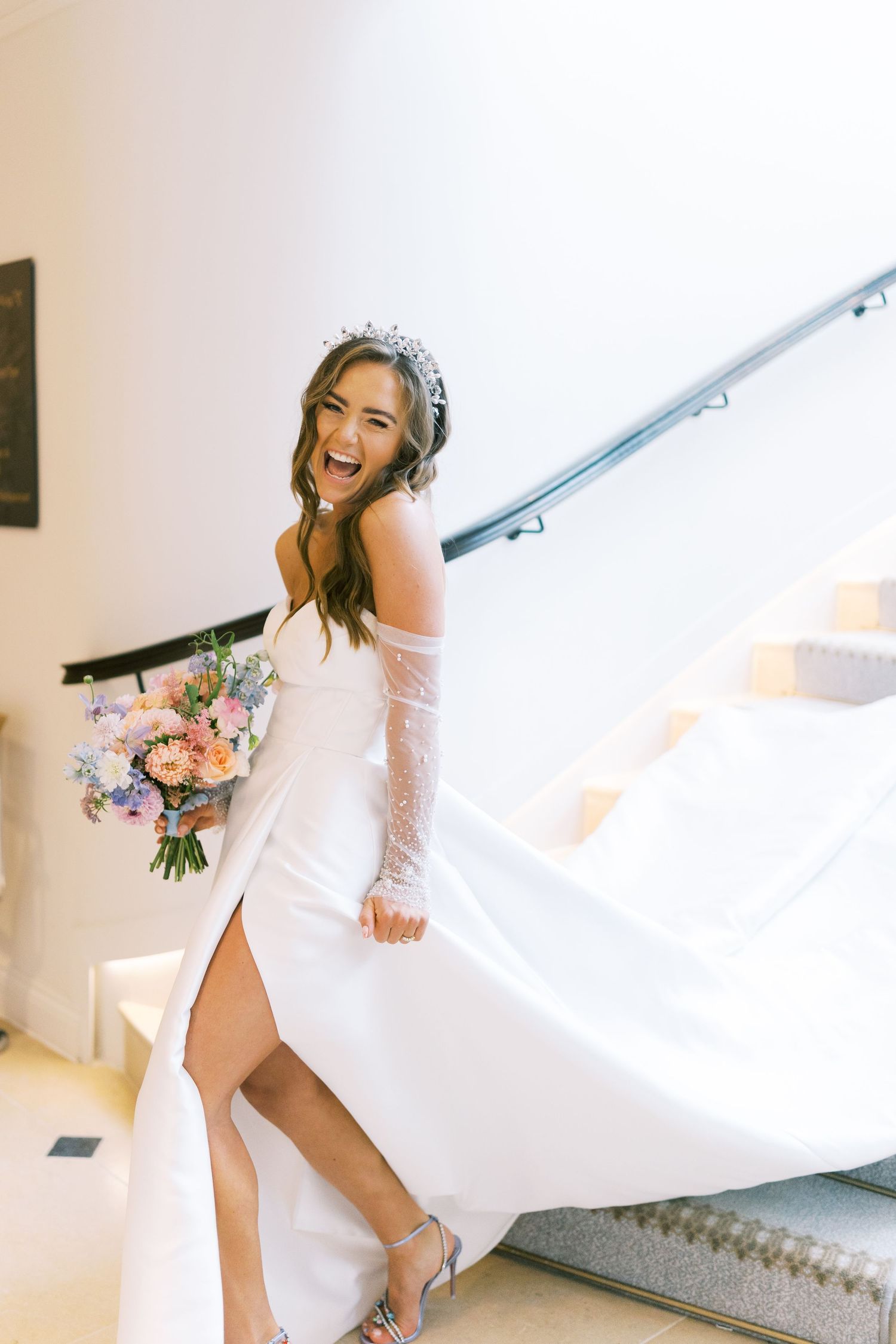 Radiant bride in off-shoulder lace wedding gown and tiara holds colorful bouquet on elegant staircase.