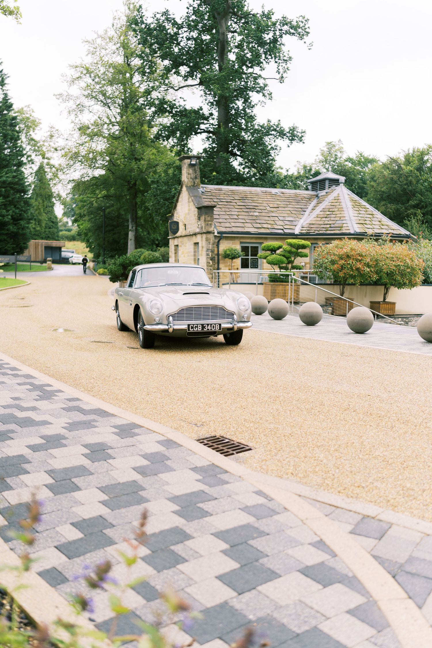 A classic silver Aston Martin DB5 parked on a gravel driveway outside a charming stone country house venue.