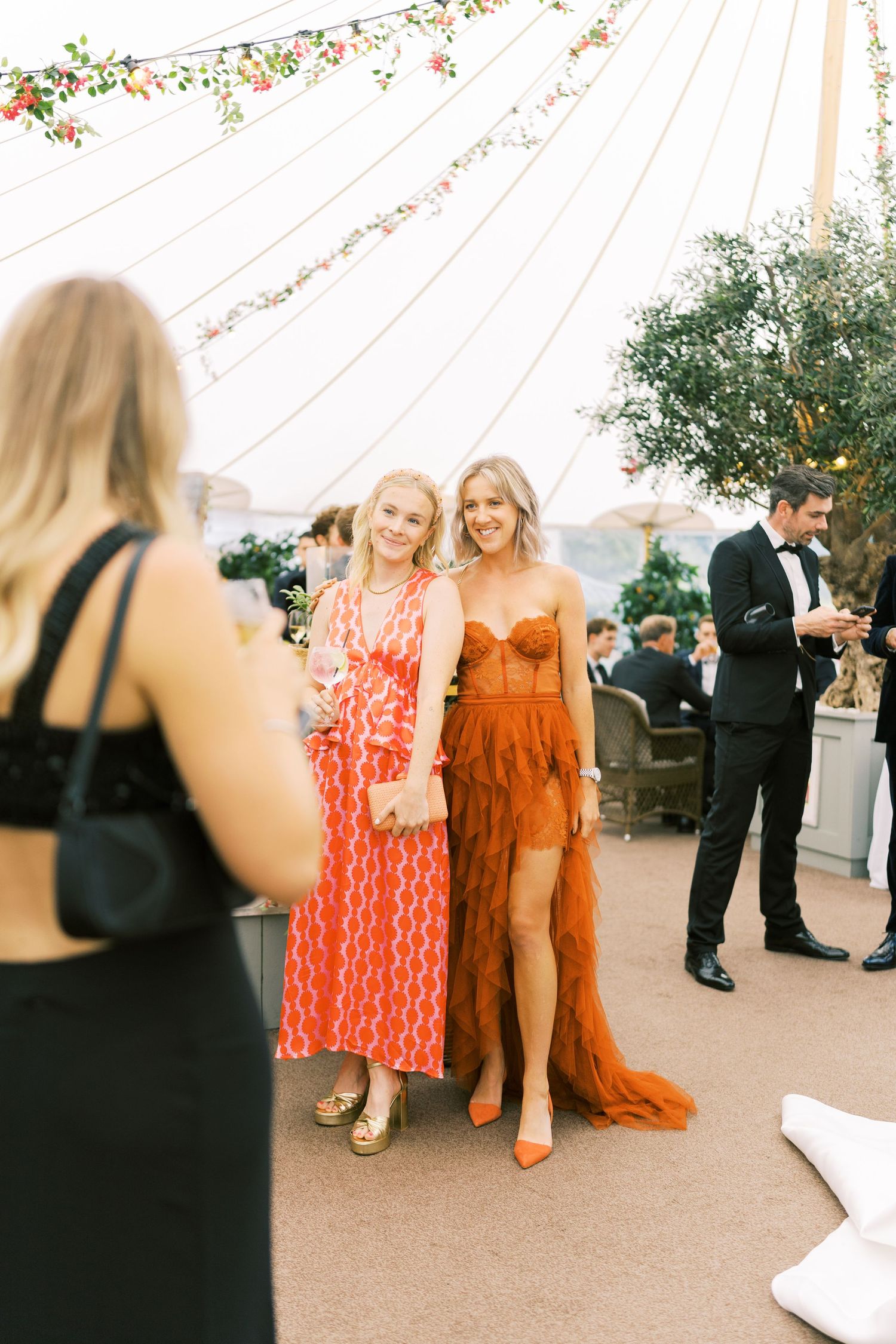 Two female wedding guests posing in red and rust strapless gowns under floral decorated tent canopy.