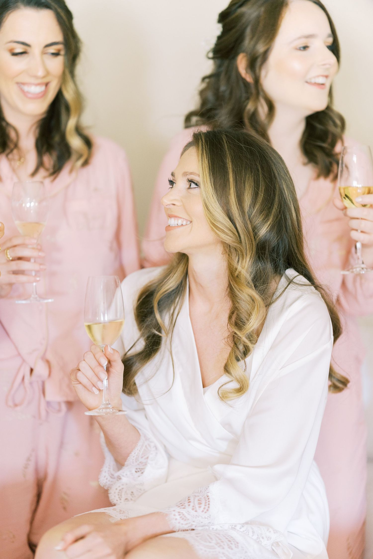 Bride in white lace robe holding champagne, smiling with bridesmaids in pink robes behind her.