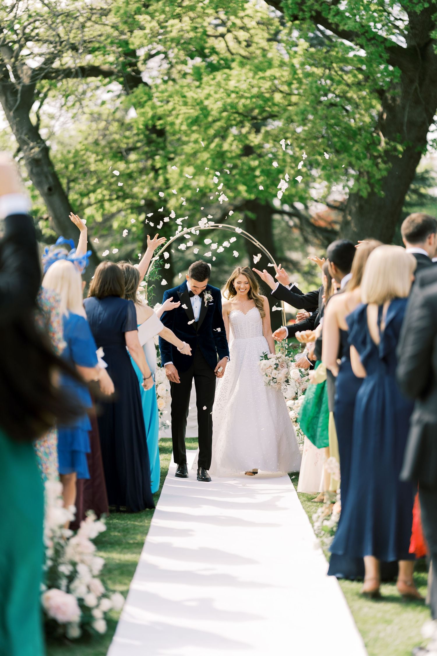 Bride and groom walk down outdoor aisle as guests toss flower petals under a circular floral arch.