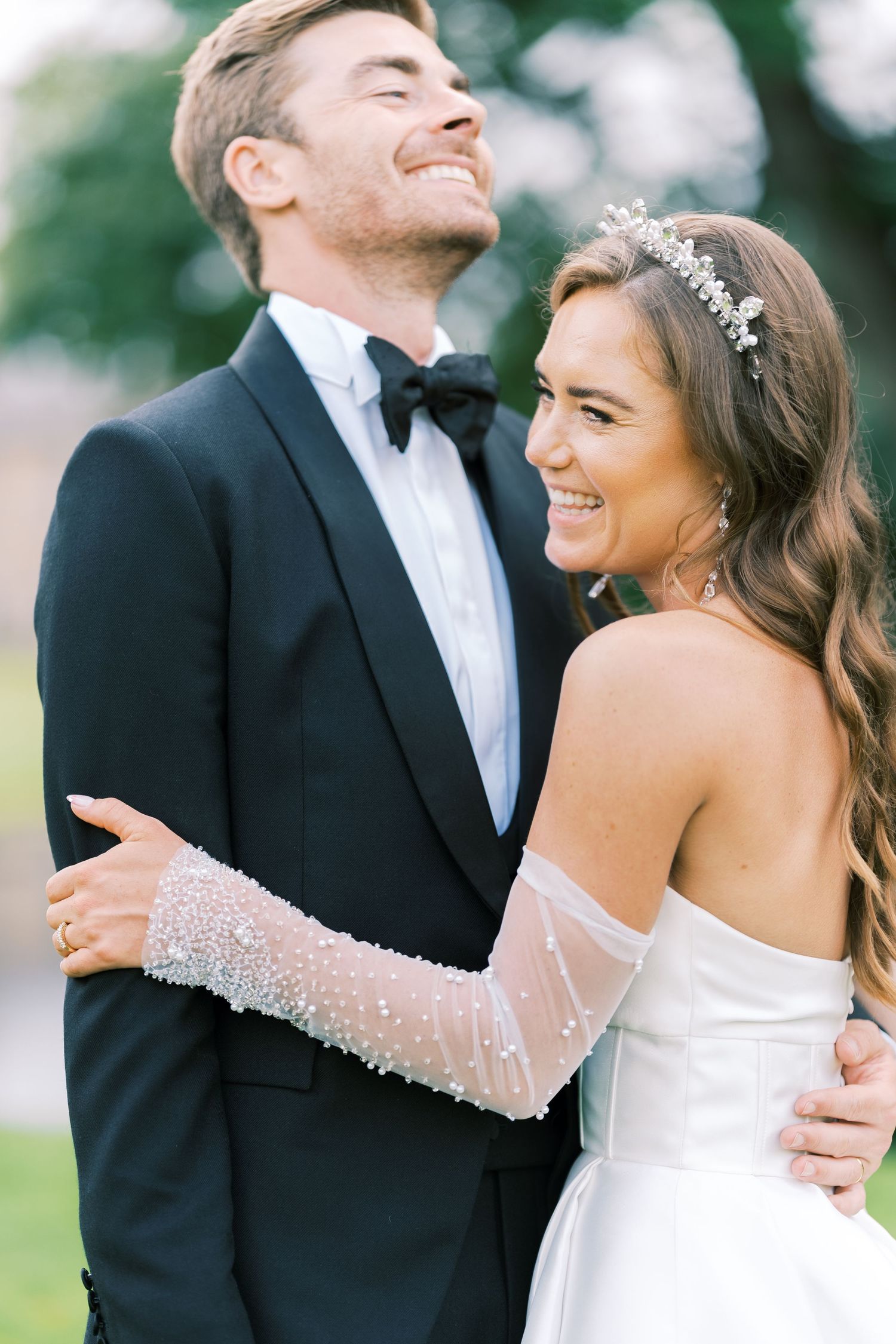 Smiling bride in off-shoulder gown with beaded sleeves and tiara embraces laughing groom in black tuxedo outdoors.