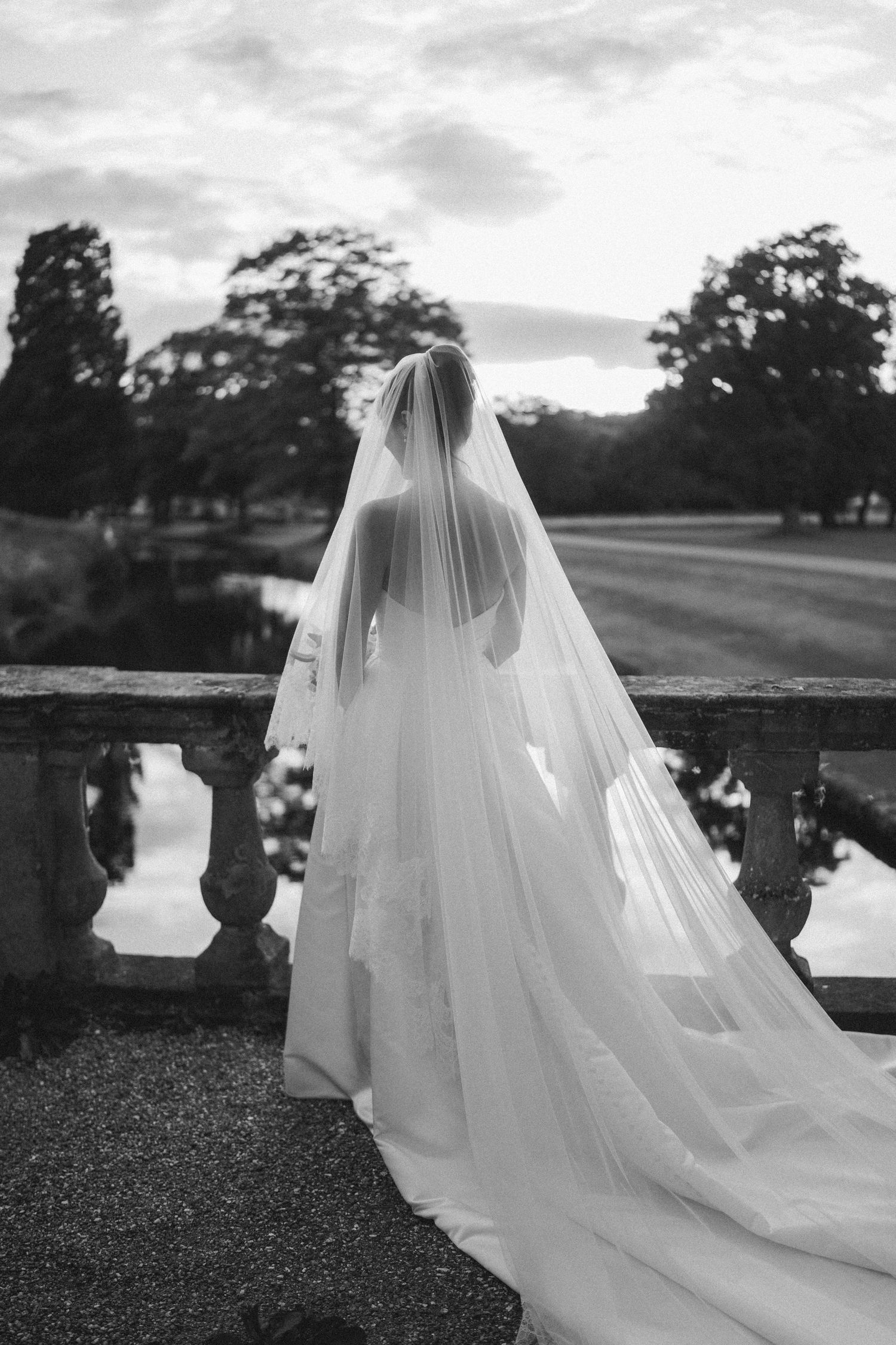 Black and white photo of a bride in a flowing gown and veil standing on a stone terrace overlooking a serene garden.