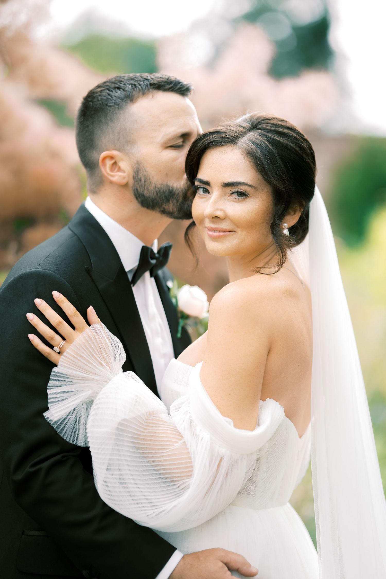 Groom in black tuxedo kissing bride in off-shoulder white gown with veil outdoors among pink blossoms.