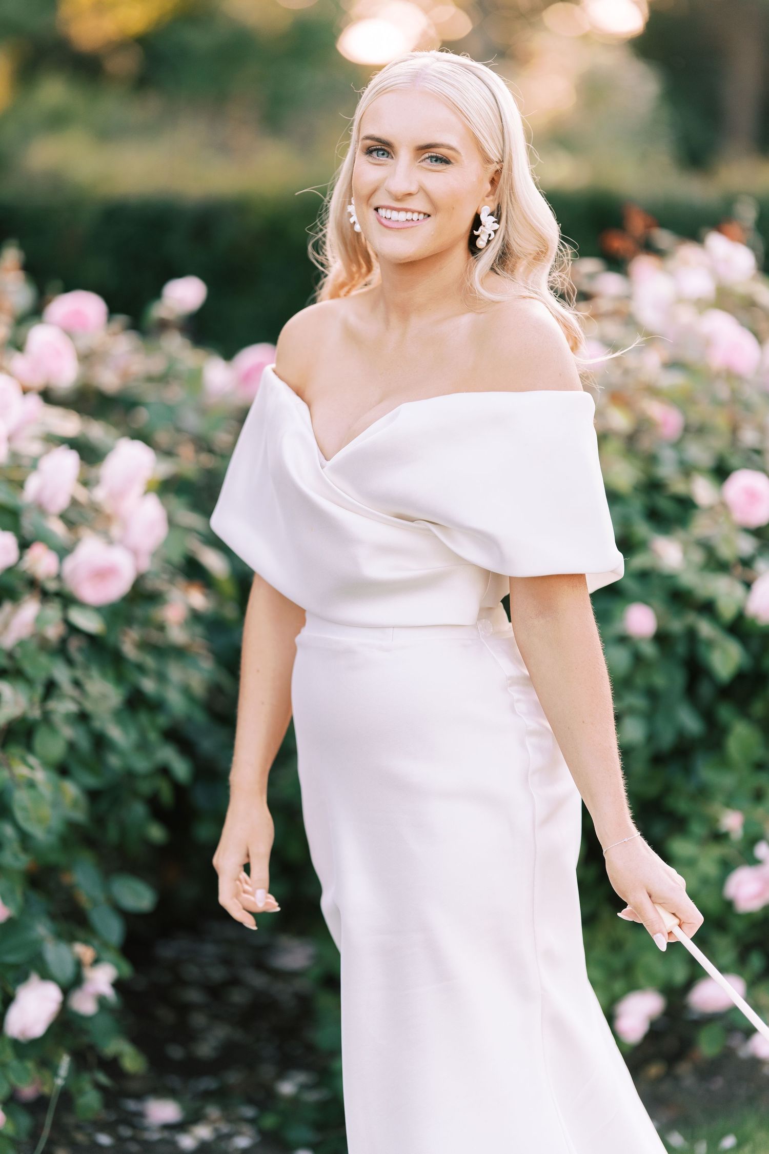 Blonde bride in off-shoulder white gown smiling among pink roses in garden setting.