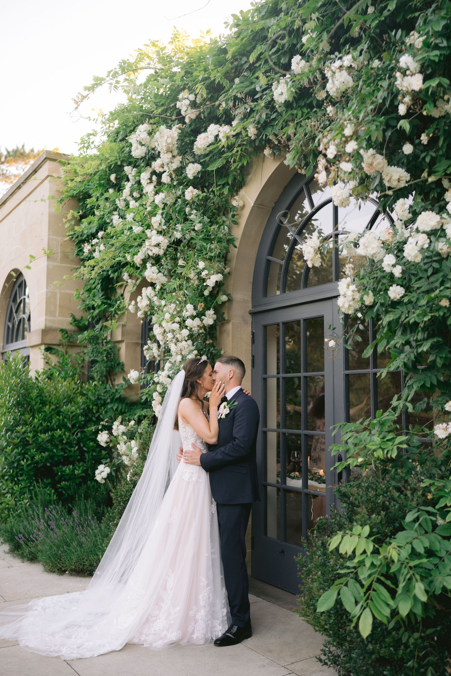 Bride and groom kissing under a floral arch of white roses on a stone building on their wedding day.