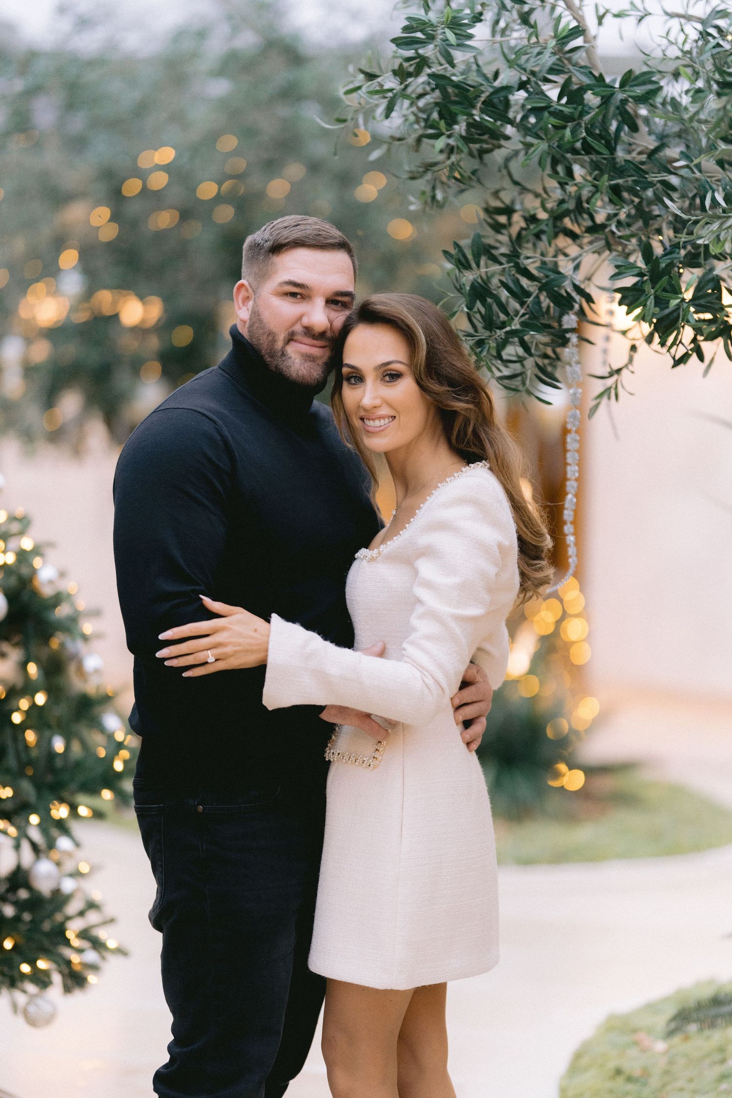 A couple embraces warmly outdoors, man in black turtleneck, woman in white mini dress, with festive lights in background.