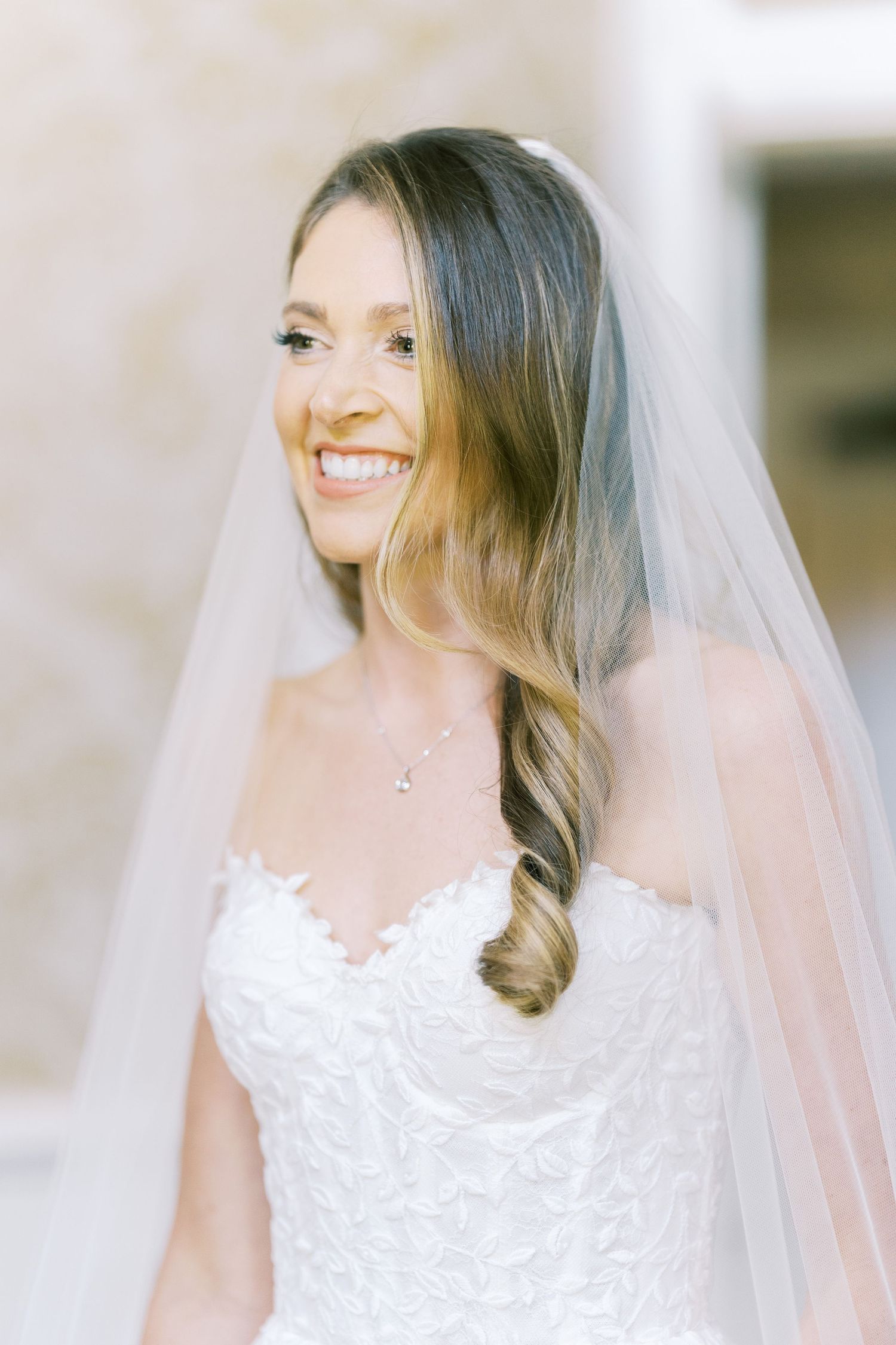 Smiling bride wearing a white lace strapless gown and veil with long wavy hair.